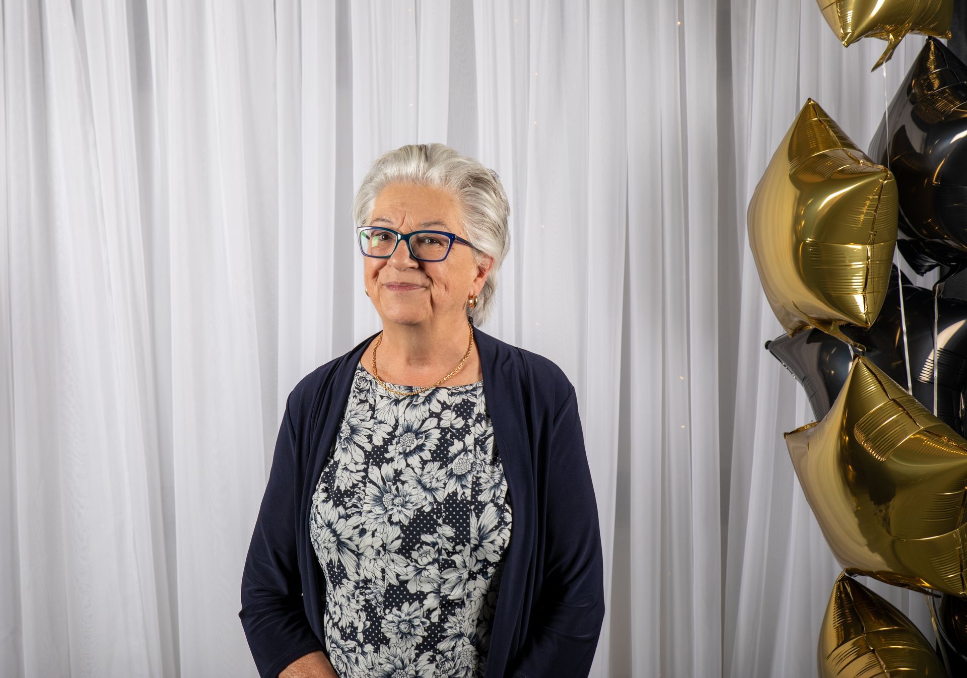 Woman with glasses smiles, standing in front of white curtains and star-shaped gold and black balloons.