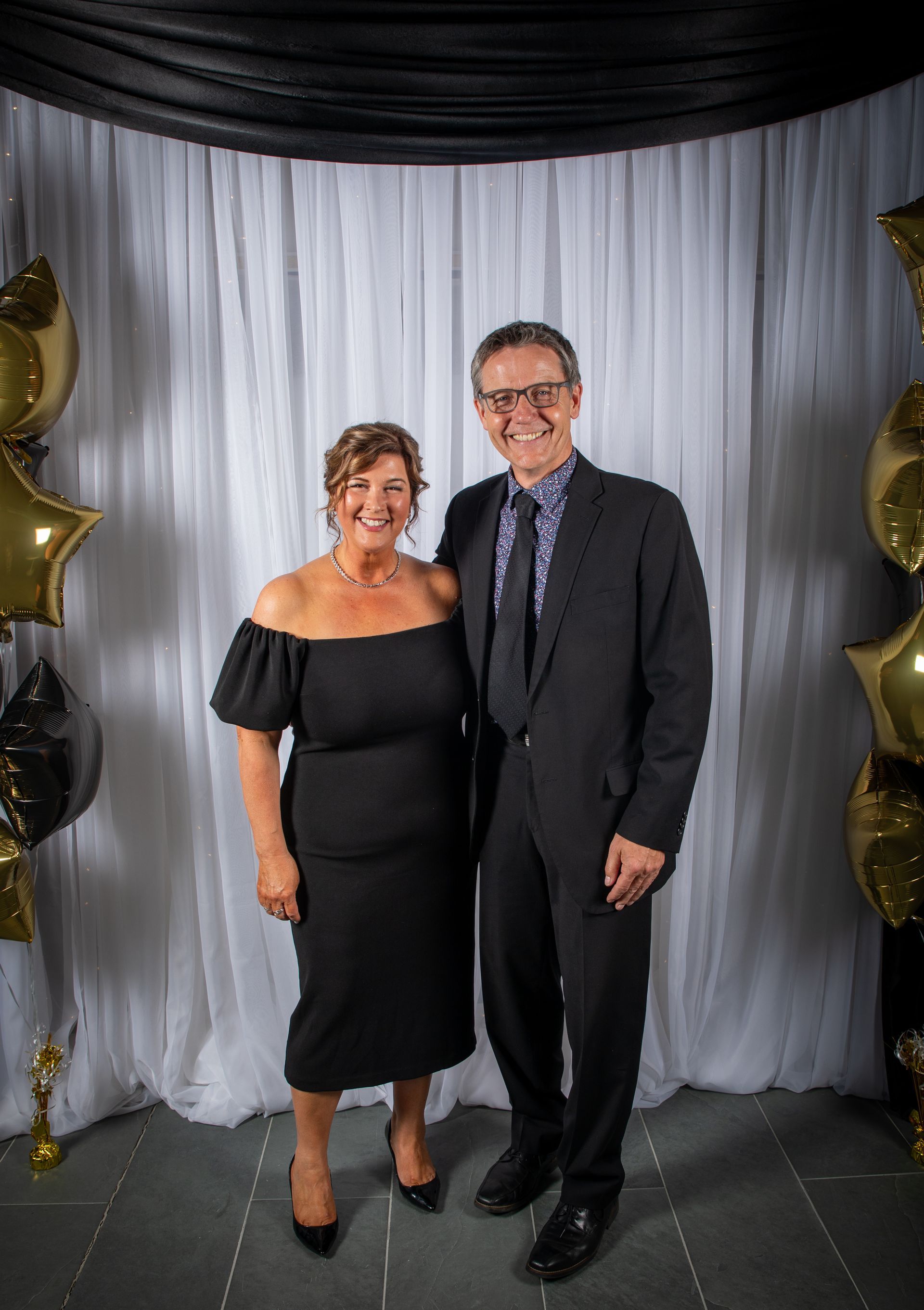 Couple in formal attire poses in front of a white backdrop with balloons.