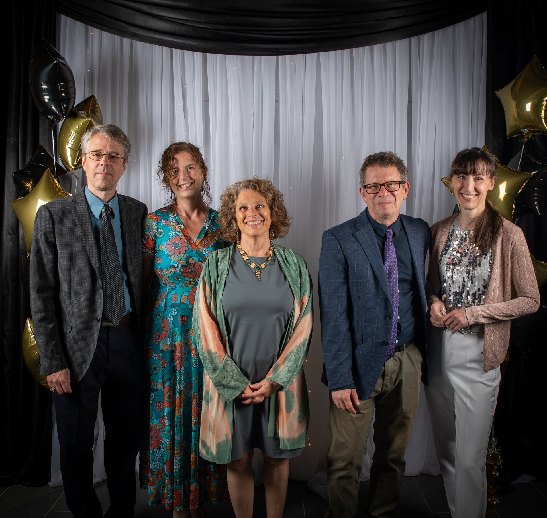 Group of five smiling people posing in front of a draped backdrop with balloons.