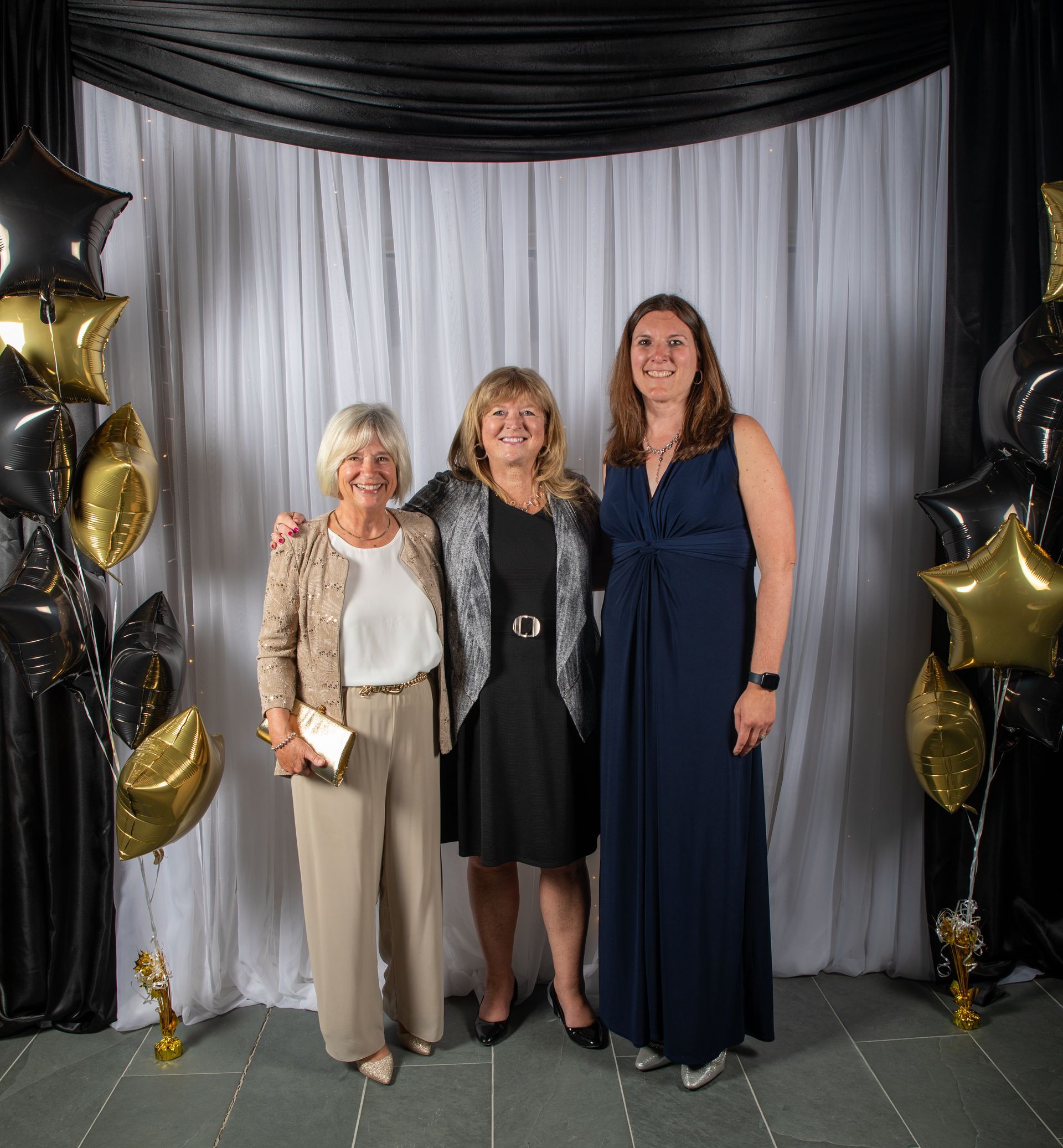 Three women pose for a photo at an event, smiling. They stand in front of a white backdrop with balloons.