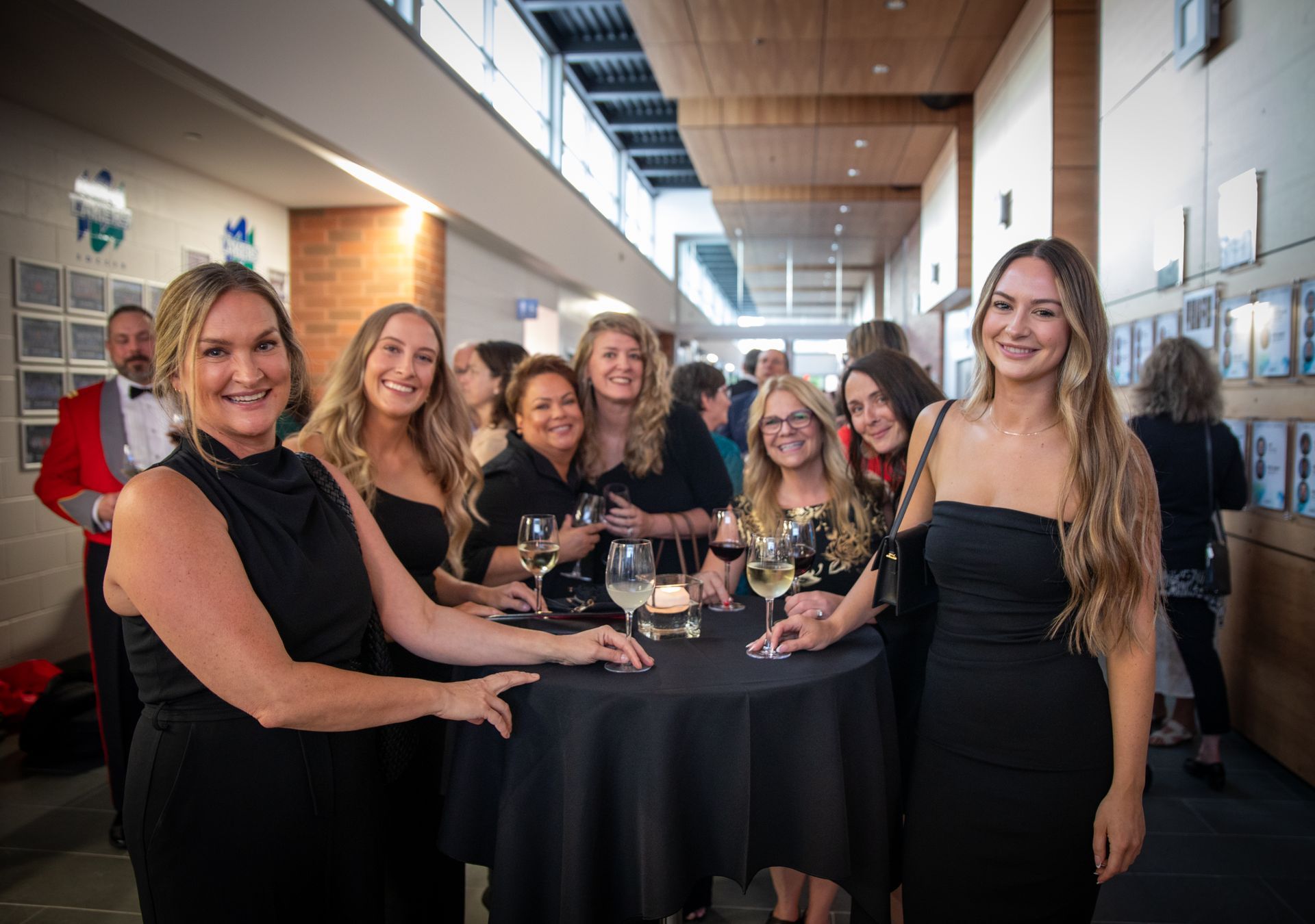 Group of women in black dresses at an event, gathered around a table with drinks, smiling.