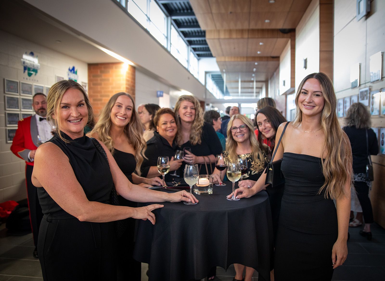 Group of women in black dresses smile at a formal event, gathered around a small table with drinks.