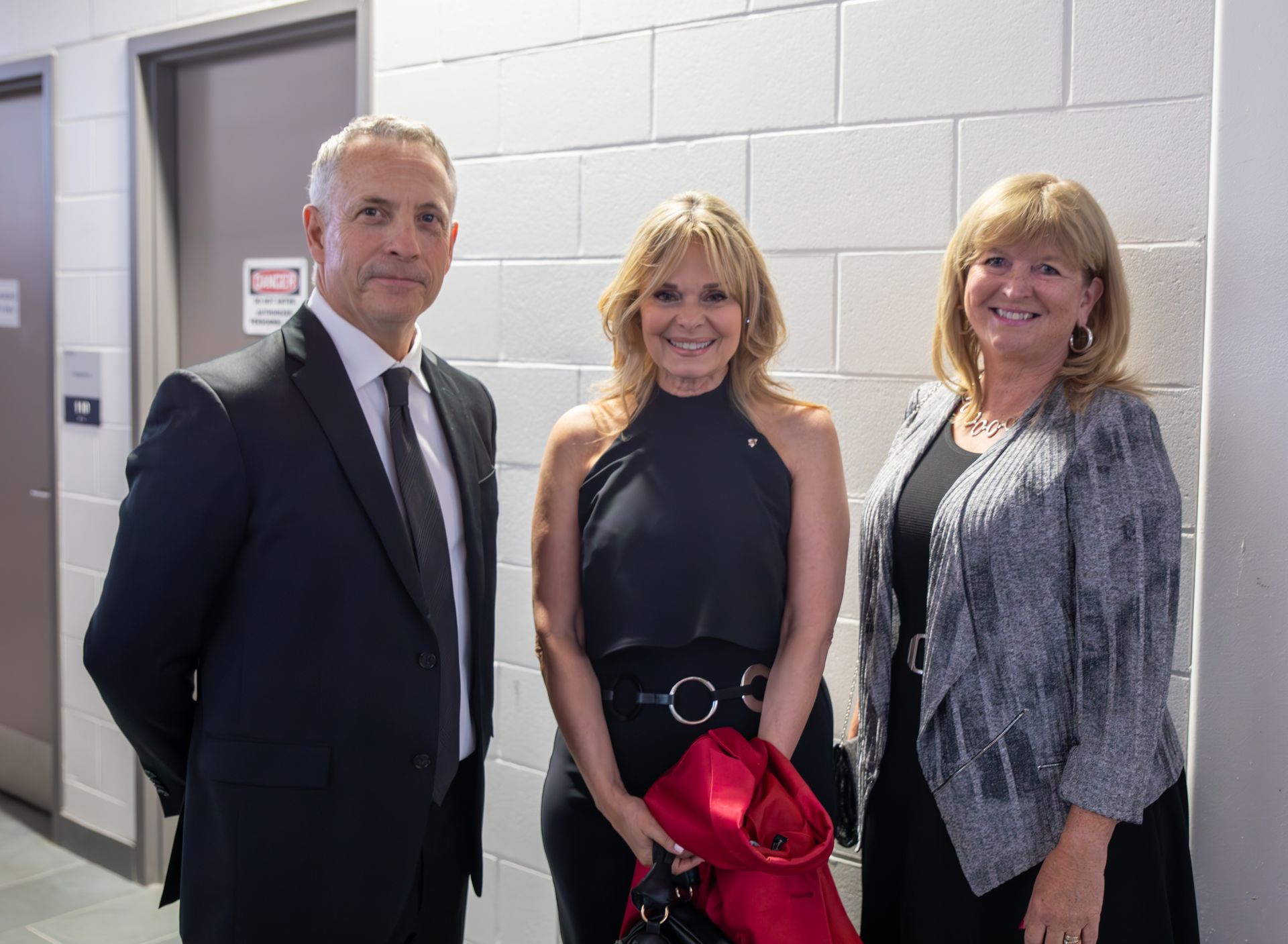 Three people standing in a hallway: a man in a suit, two women in evening attire, smiling.