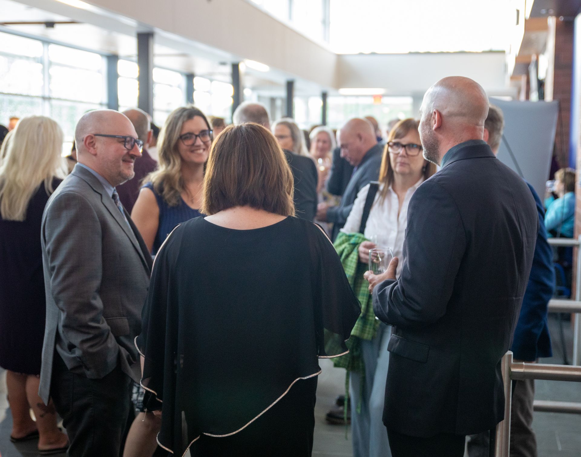 People socializing at an event, indoors. Several are in small groups, talking and smiling. Light-filled space.