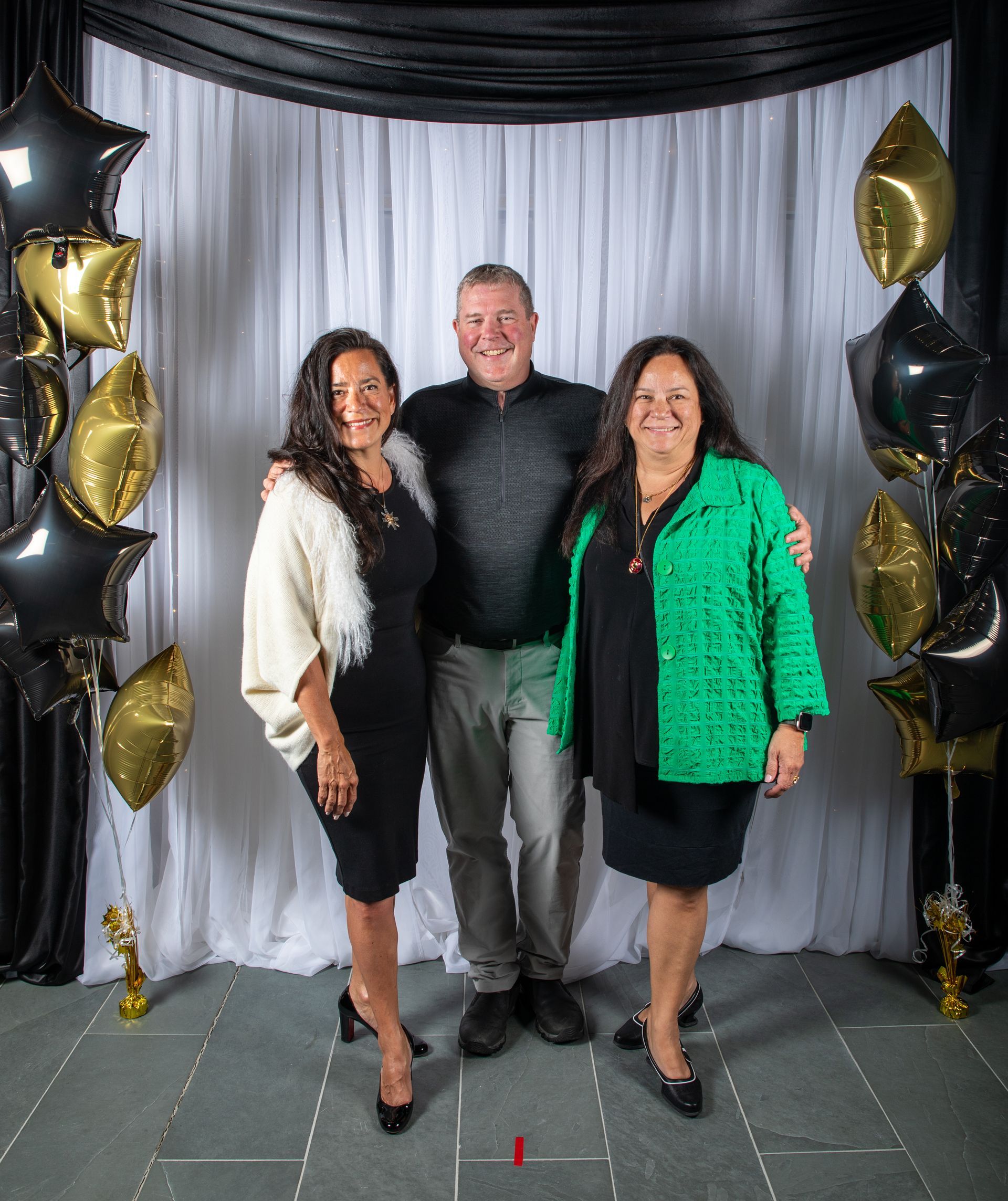 Three people posing for photo. Man in center, two women on either side. Black, gold balloons.