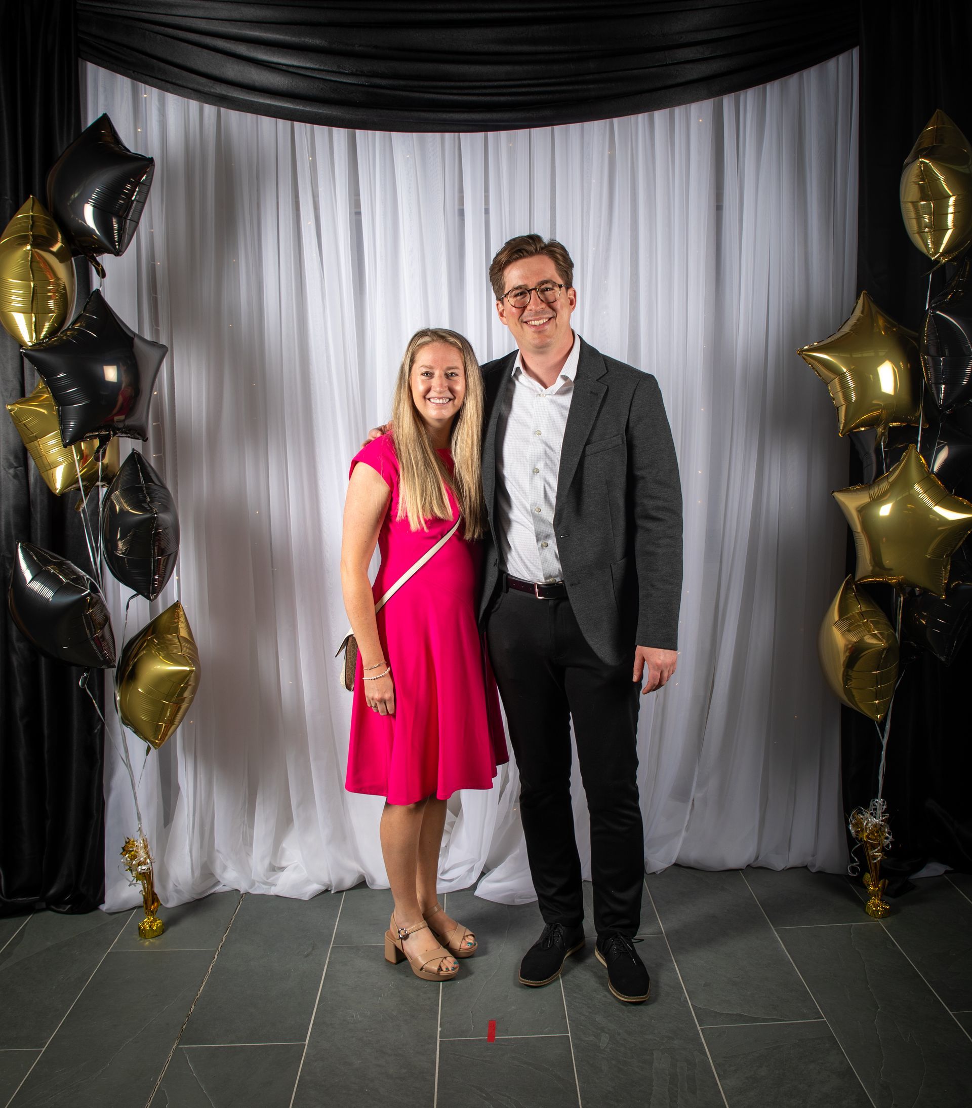 A woman in a pink dress and a man in a suit pose together at a formal event with gold and black balloons.