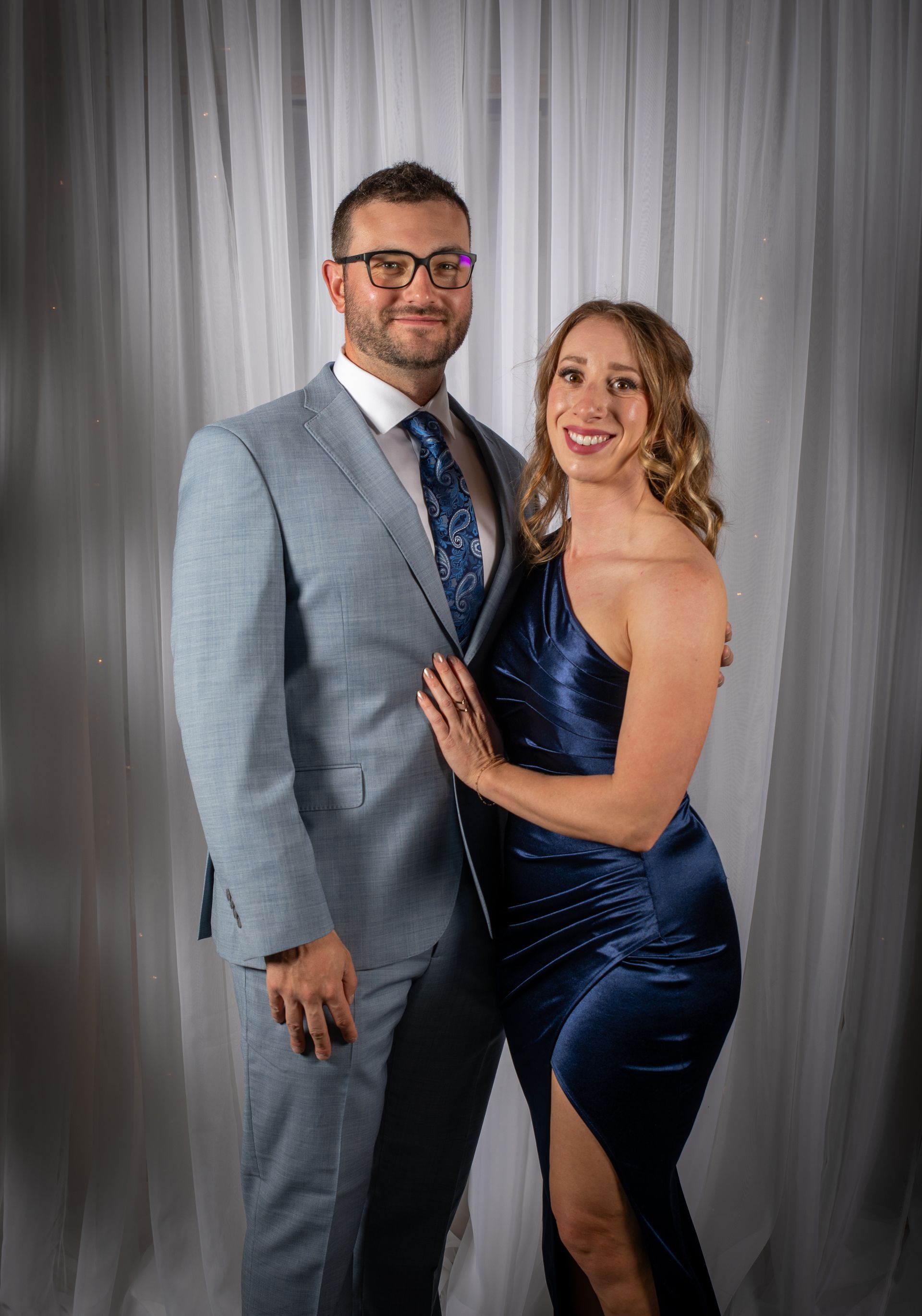 Man in a blue suit and woman in a navy gown, smiling, posing together against a sheer curtain backdrop.