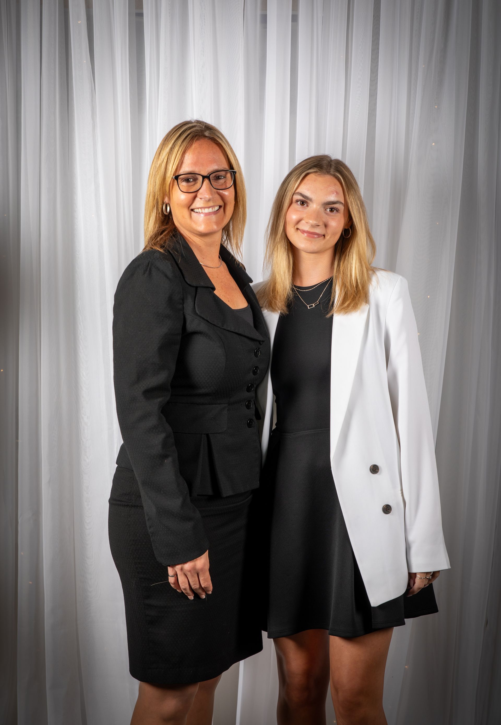 Two women pose in front of white curtains. One wears a black suit, the other a black dress with a white blazer.