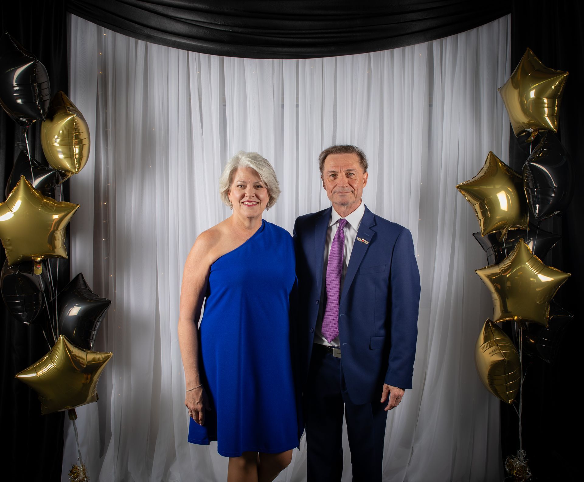 Woman in blue dress and man in suit pose in front of a curtain backdrop with gold and black star balloons.