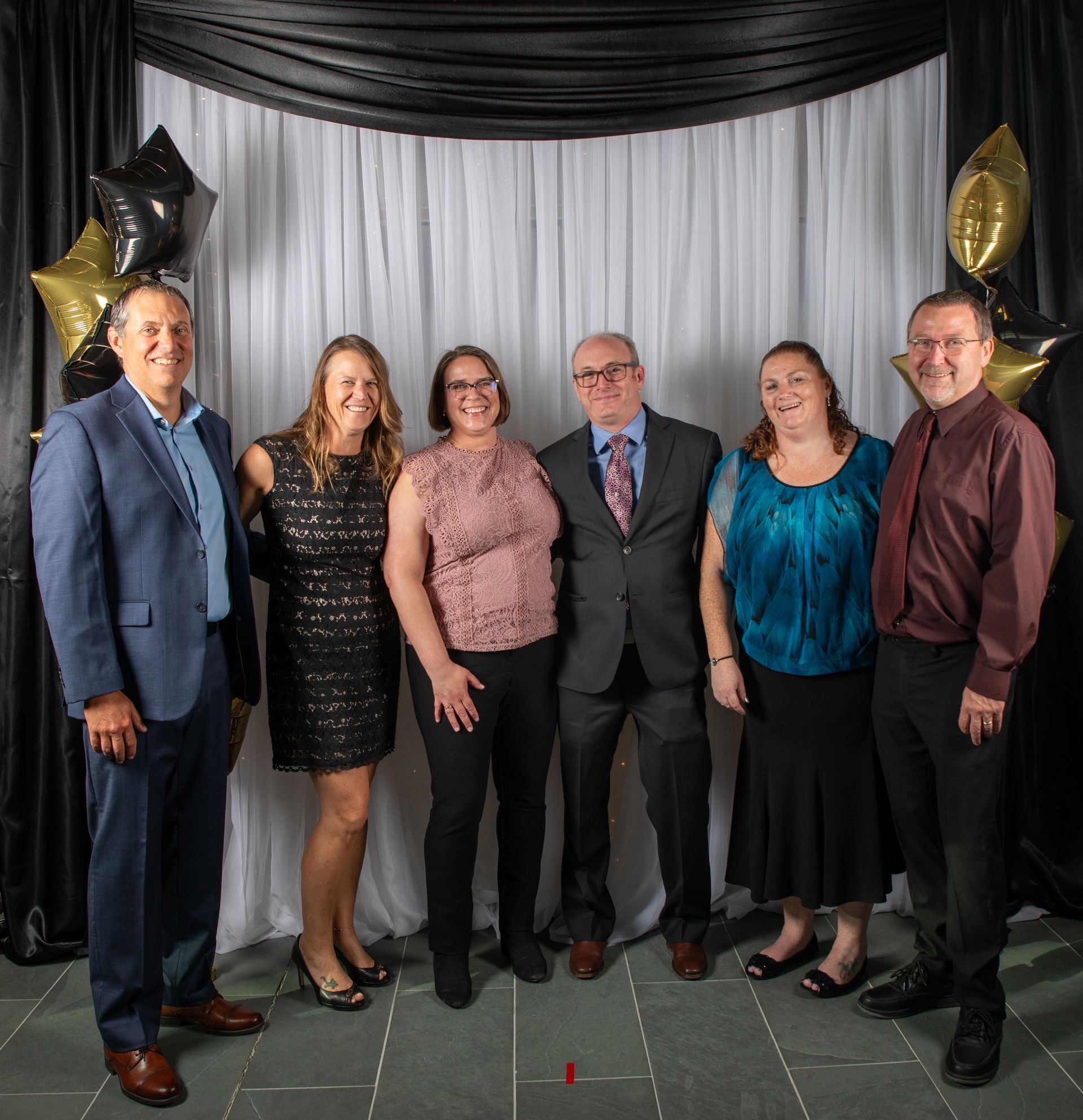 Group of six people posing for a photo. White backdrop, balloons, formal attire.
