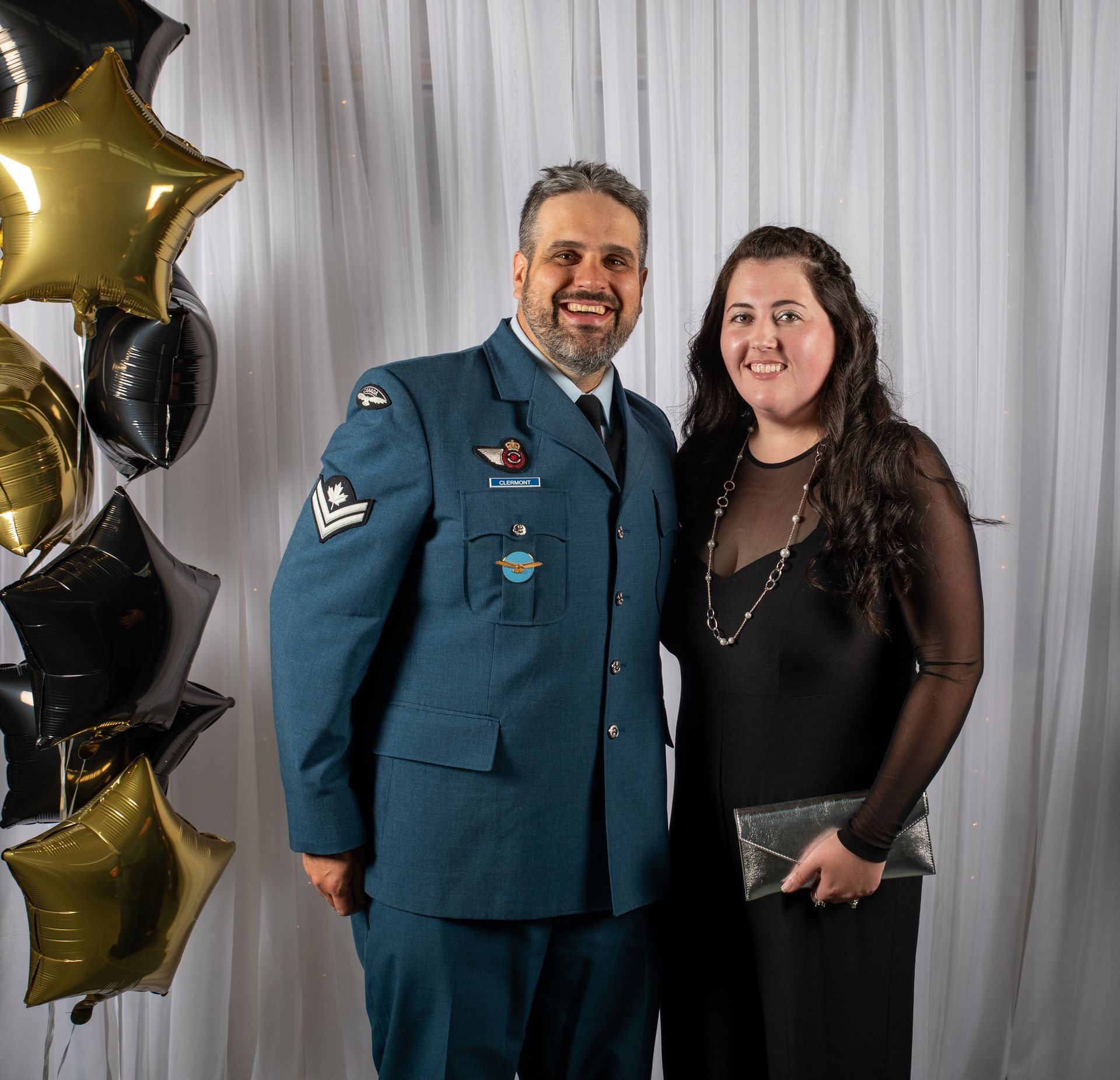 Man in uniform and woman in black dress smiling, posing together at formal event.