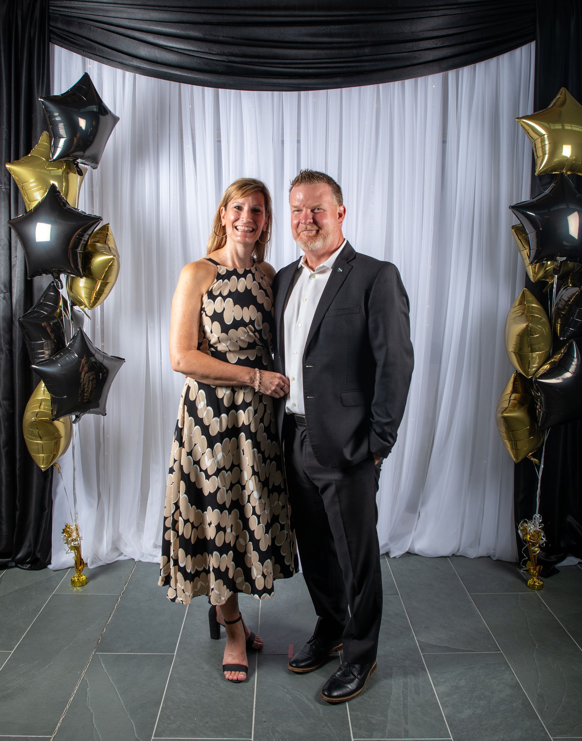 Couple posing in front of black and gold balloons and backdrop. Woman in dress, man in suit.