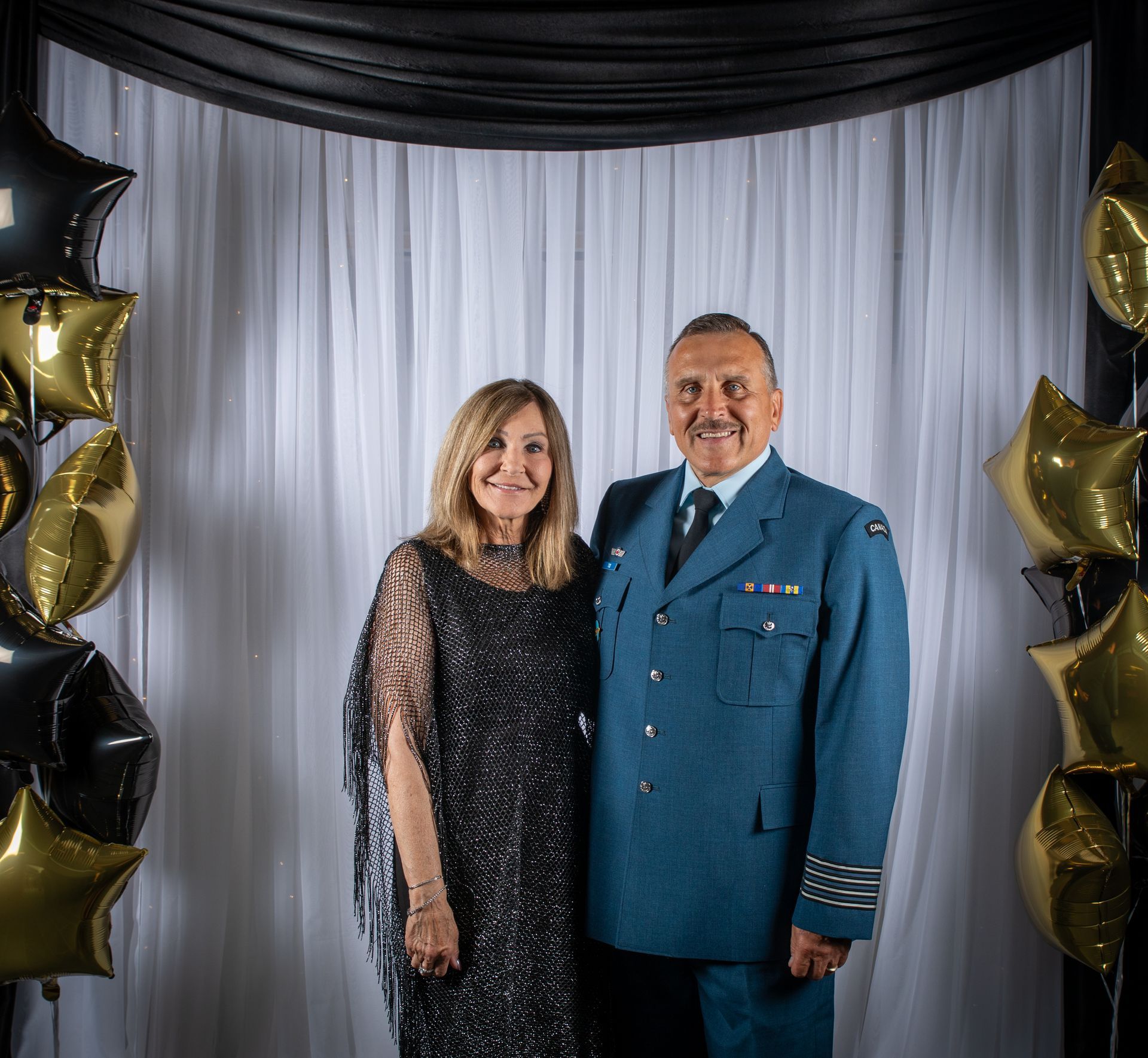 A couple poses in front of a backdrop with balloons. Man in uniform, woman in a dress.