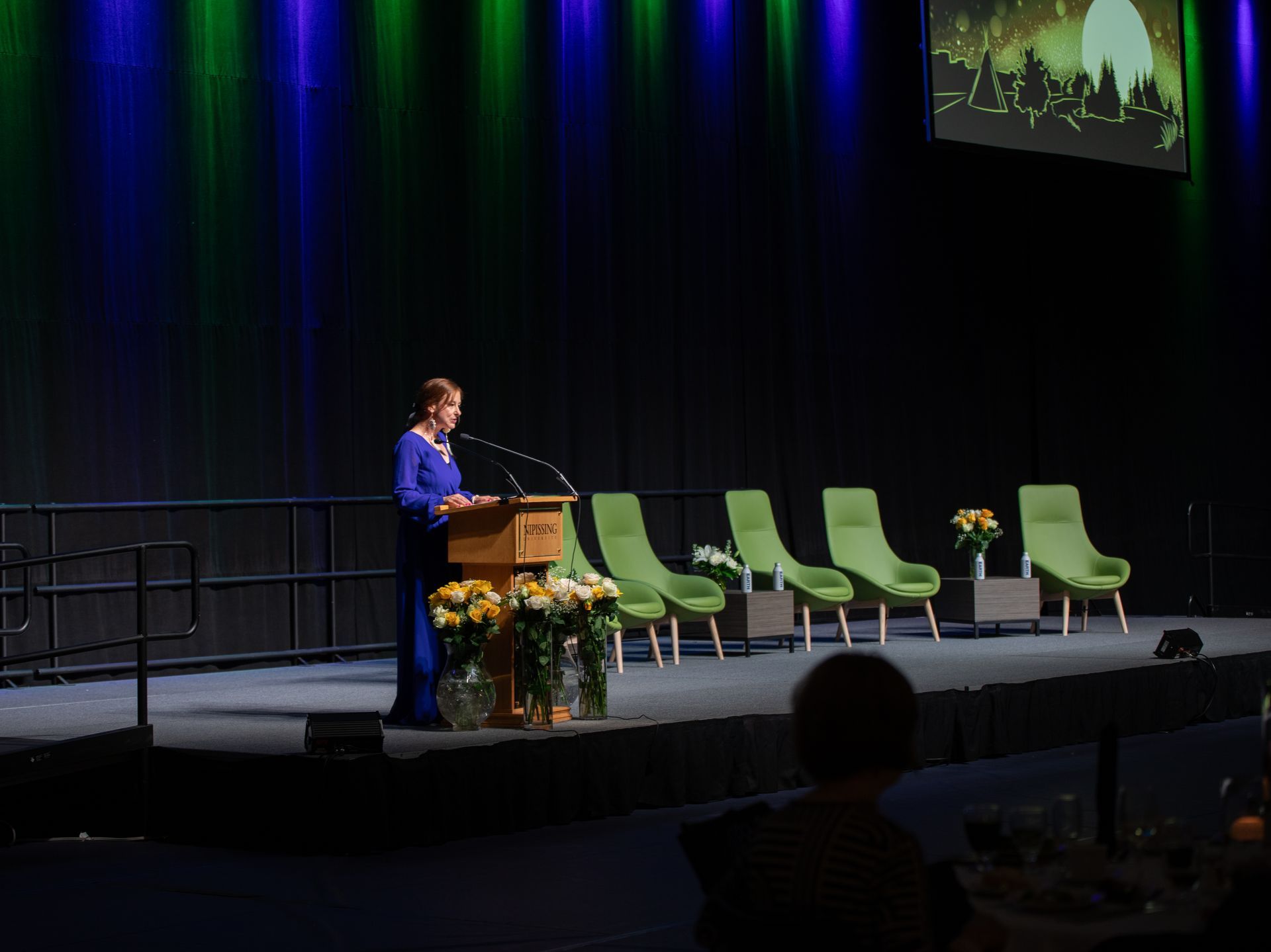Woman speaking at a podium on a stage, with green chairs and flowers. Green and blue lights in background.