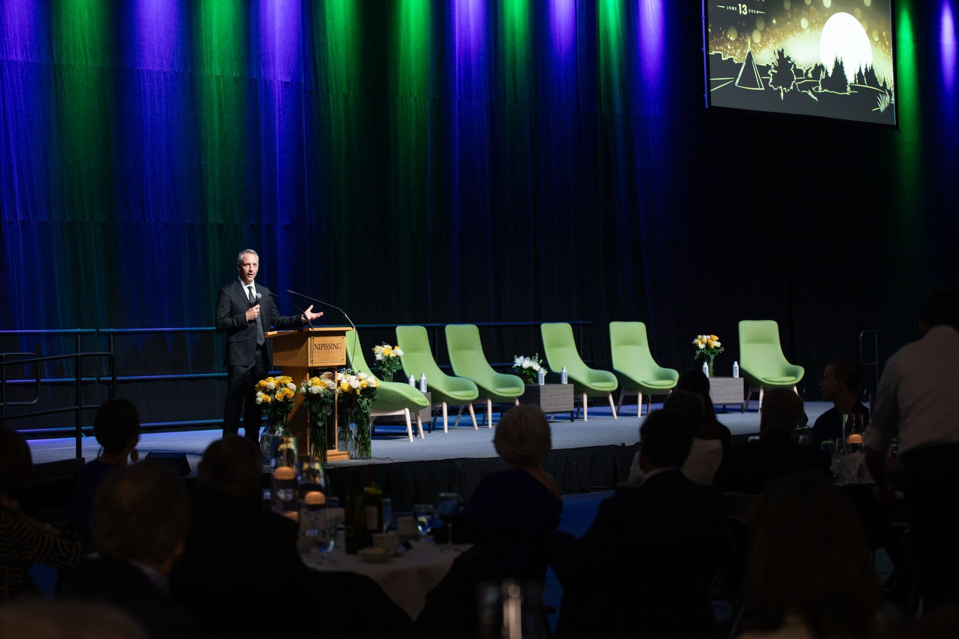 Man speaking at podium on stage with chairs, audience seated in front, stage lit with blue and green.