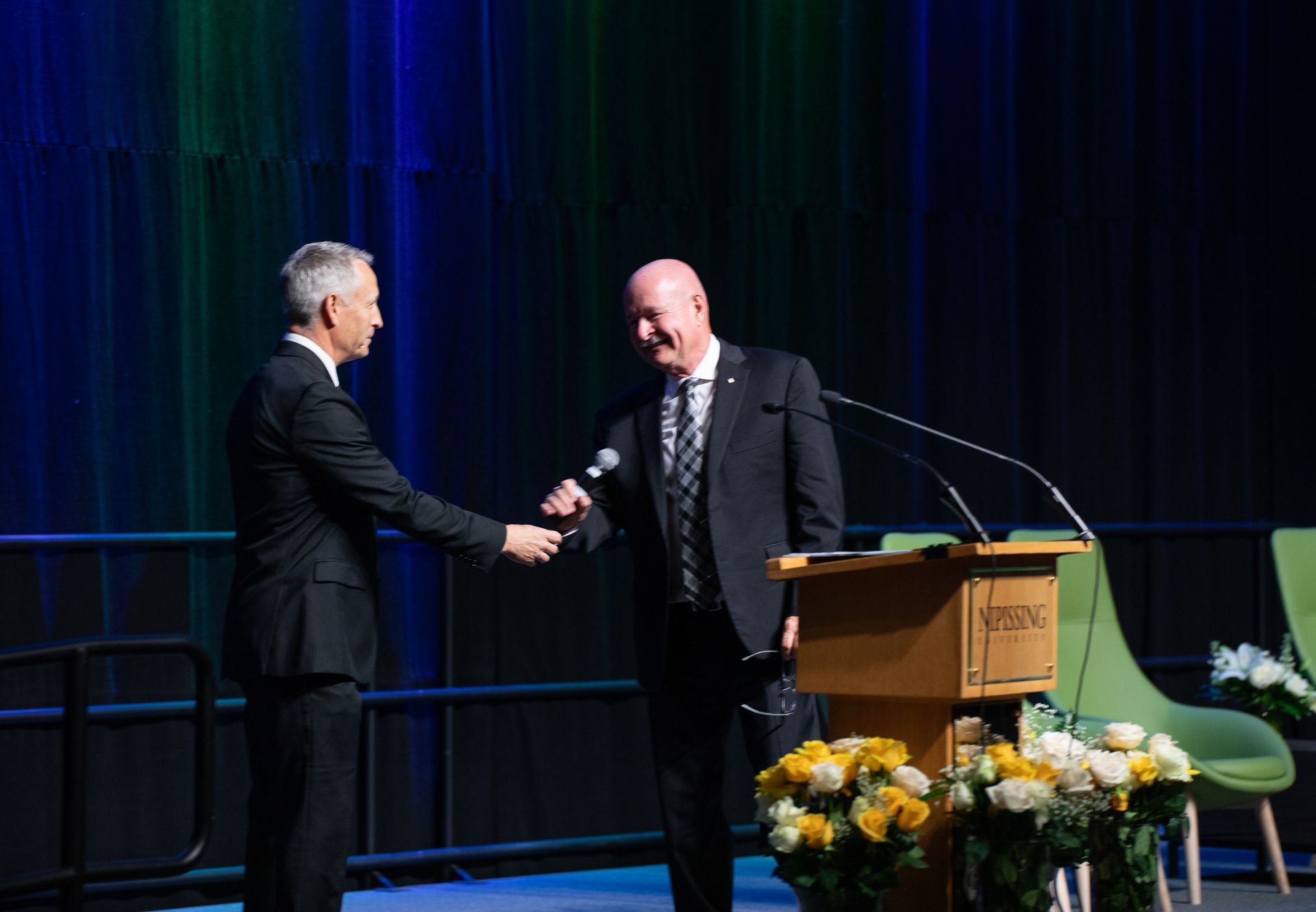 Two men in suits shake hands on a stage with a podium and flowers.