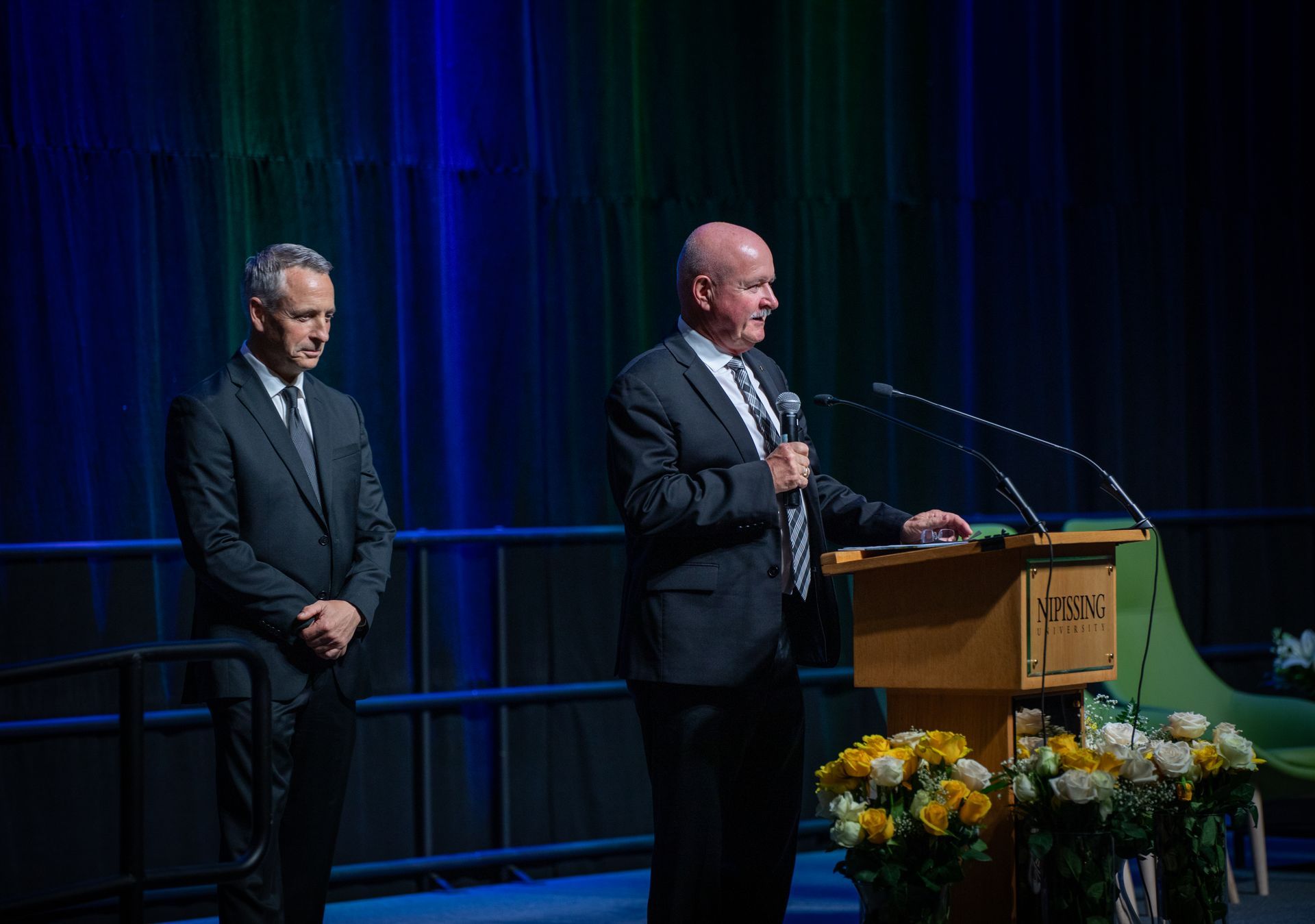 Two men in suits at a podium; one speaks into a microphone, the other stands by. Stage with flowers and blue backdrop.
