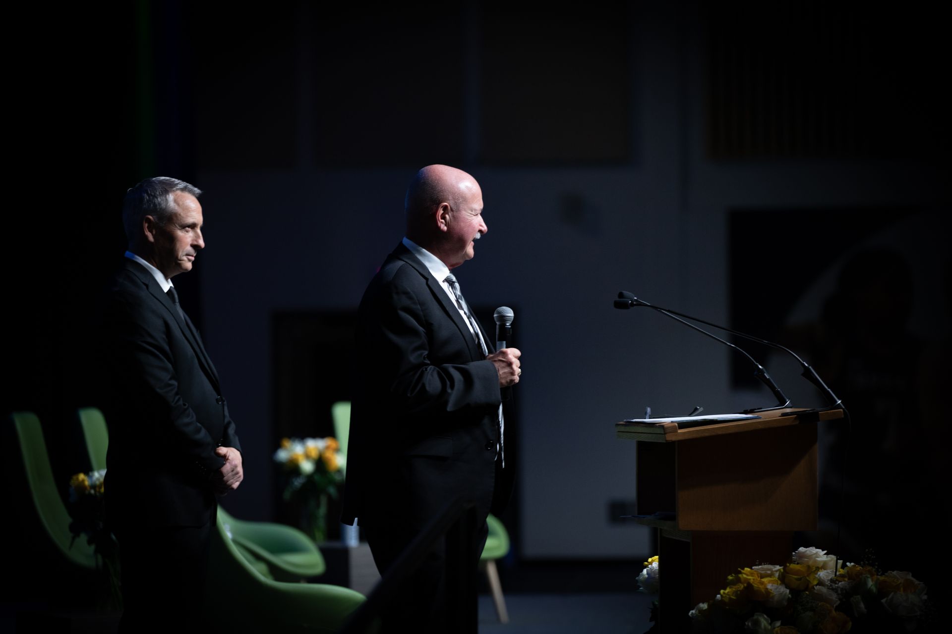 Two men in suits on stage. One speaks into a microphone, the other stands beside.