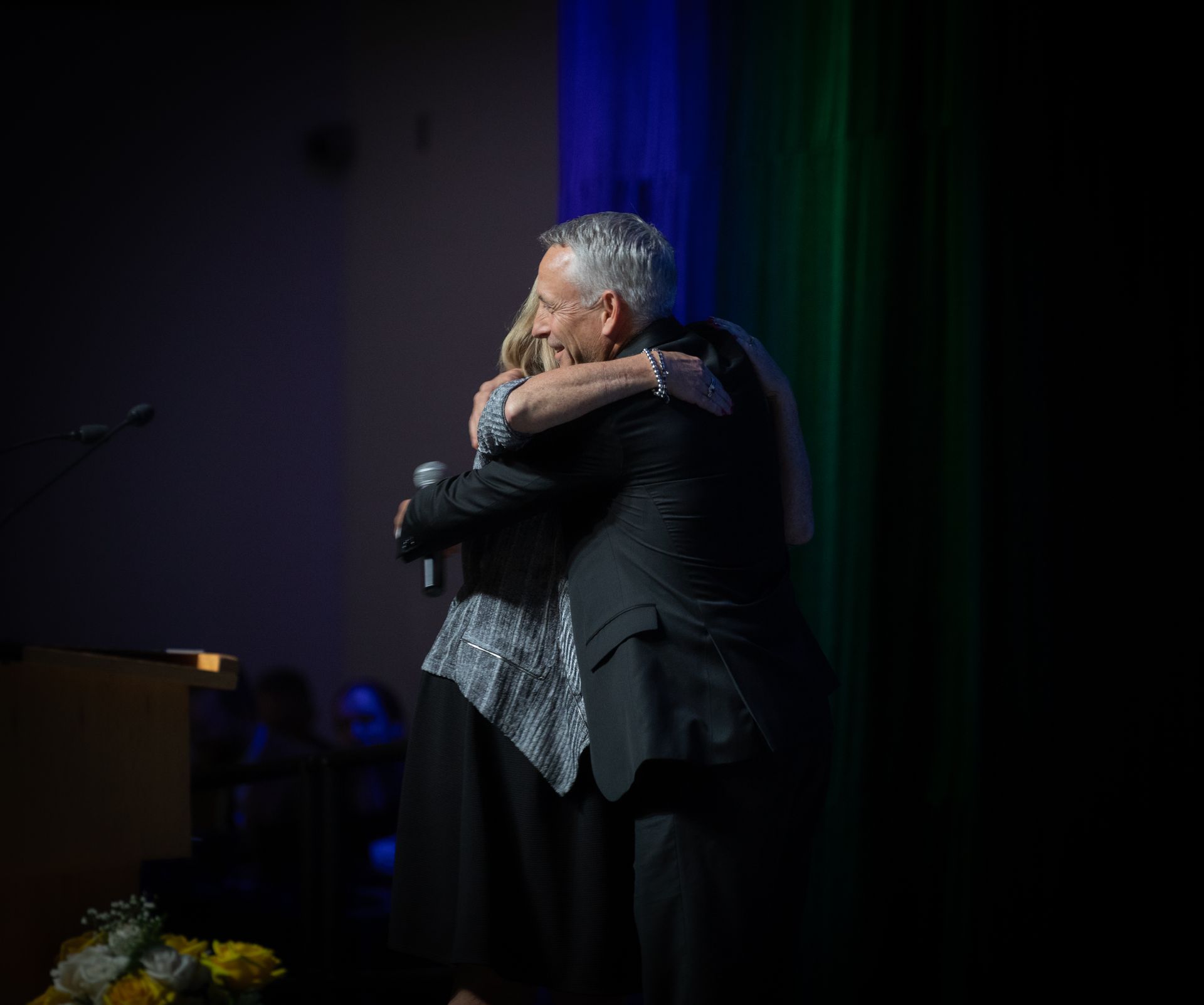 Man in suit hugs woman on stage, illuminated by stage lights, blue and green backdrop.