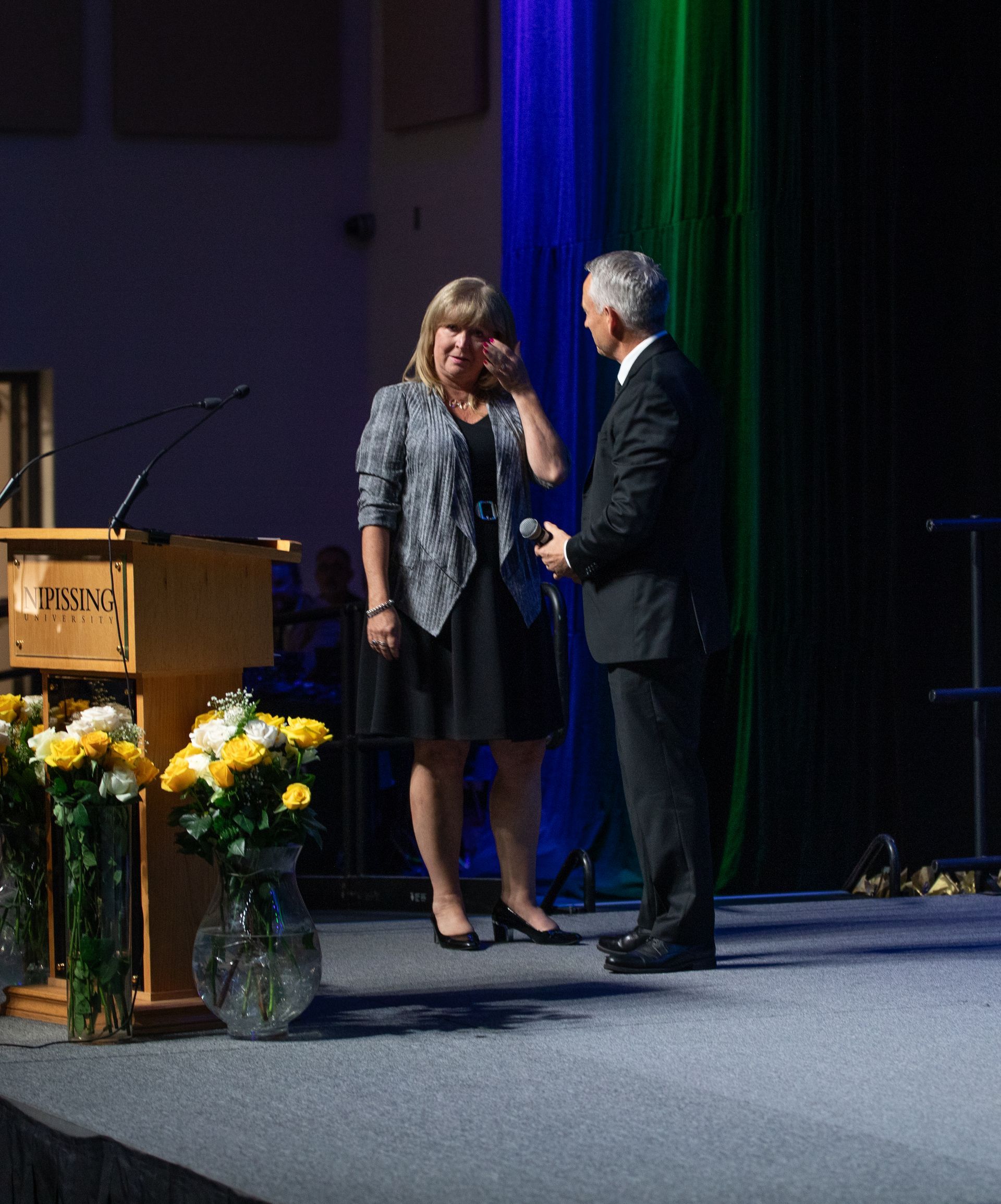 Woman wiping eye, receives award on stage, with presenter; stage backdrop, flowers.