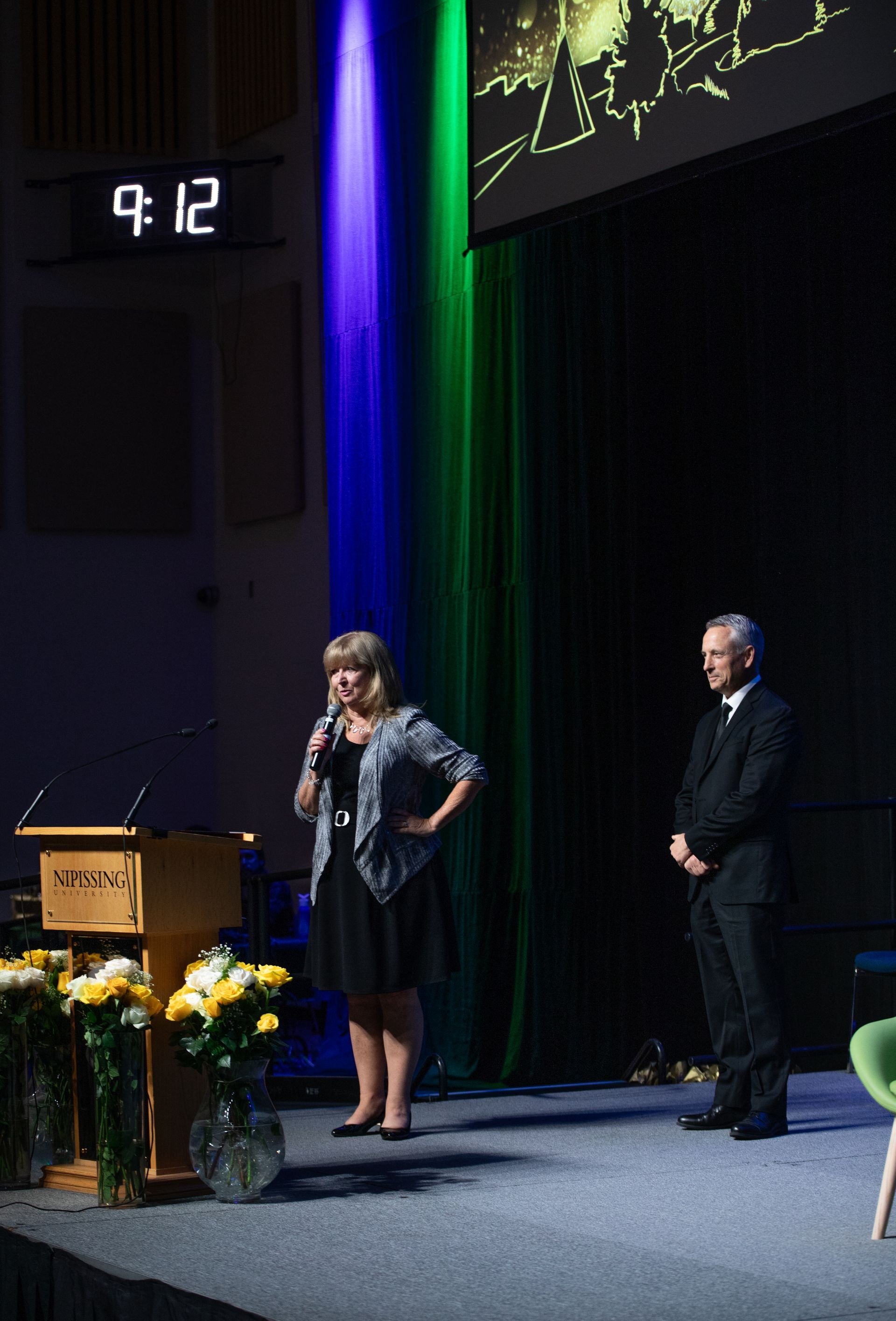 Woman speaking at a podium, man stands beside her on stage. Blue/green backdrop. 