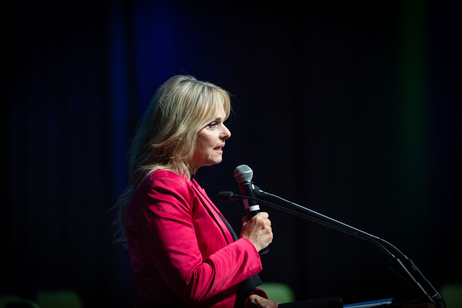 Woman in pink jacket speaking into a microphone on stage.