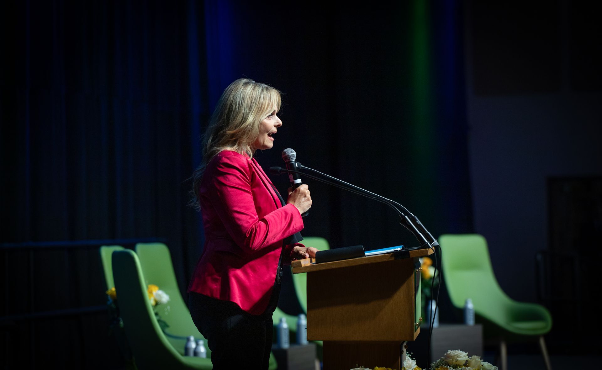 Woman in pink jacket speaks at a podium on stage; green chairs in background.