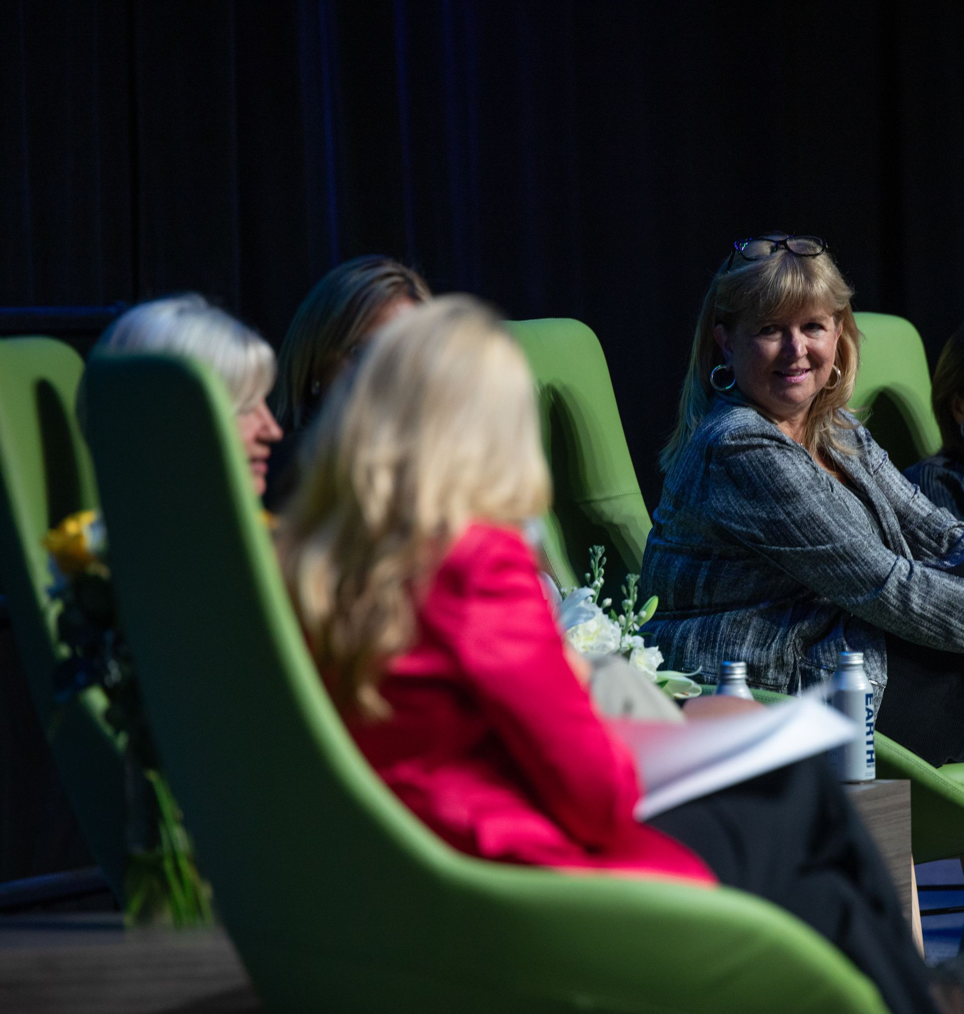 Panel of women seated on green chairs, one smiling at the camera. Dark stage background.