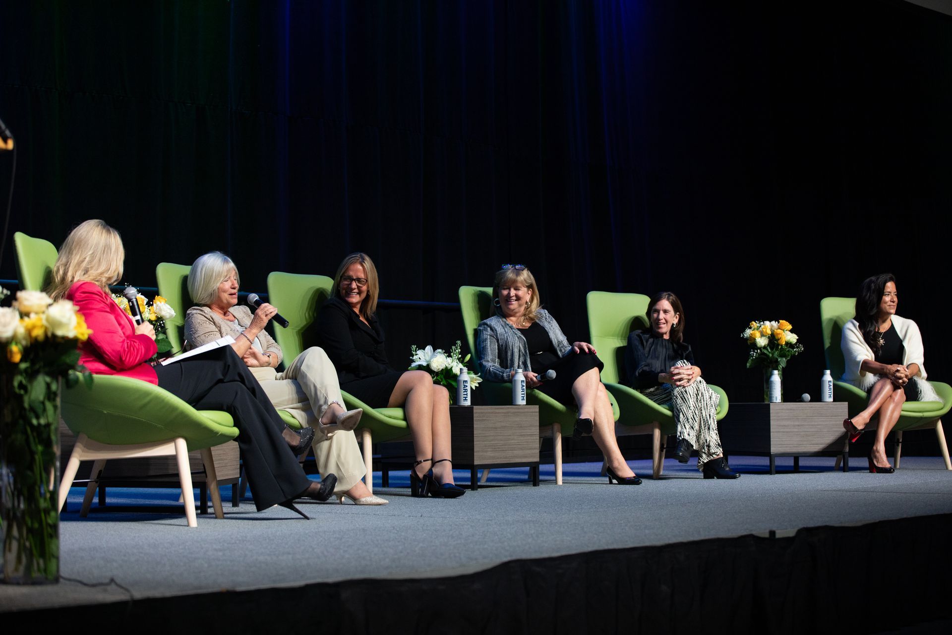 Panel of six women seated on stage, discussing. Green chairs, dark backdrop.
