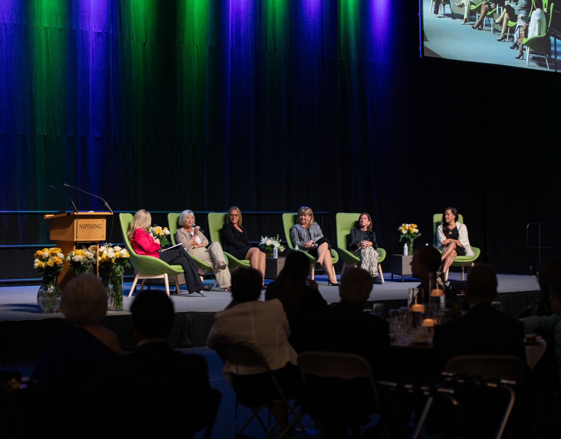 Panel of six women speaking on stage, audience in foreground. Green and blue stage lighting.