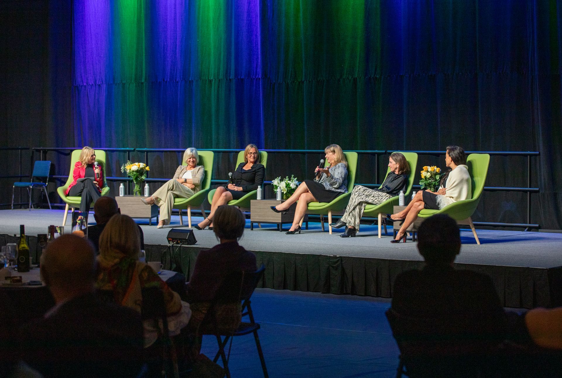Panel of six women on a stage, speaking. Audience in front. Green and blue stage backdrop.