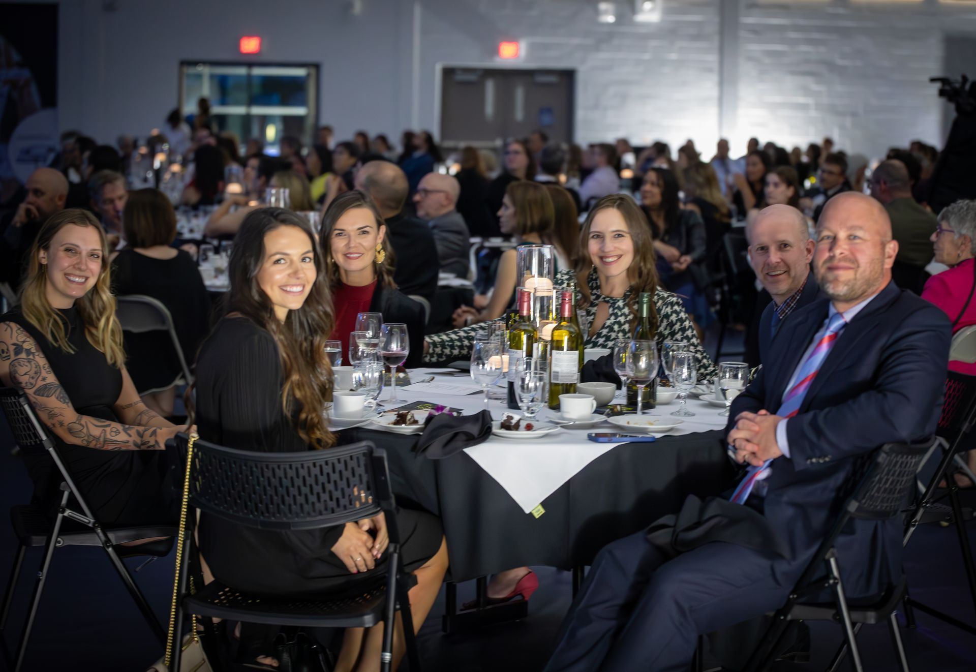 People at a gala, sitting at a table, smiling. Formal attire, indoor setting.