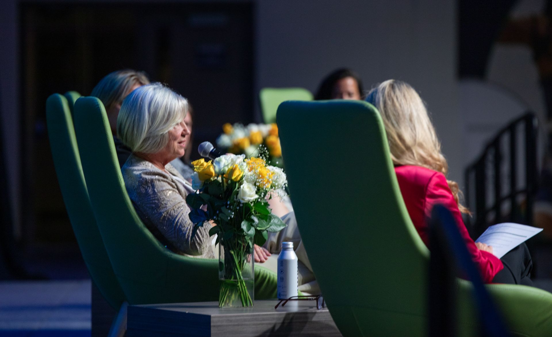 Women in green chairs at a panel discussion. Flowers on a small table.