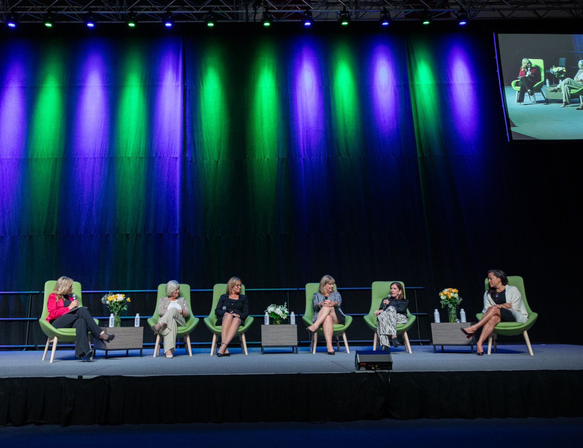 Panel of women seated on a stage, discussing. Colorful stage lighting with a screen displaying a similar view.