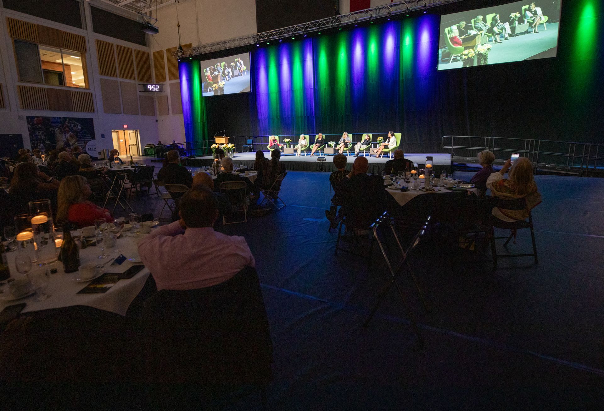Event with people seated at tables, watching speakers on a stage lit with green and blue lights.