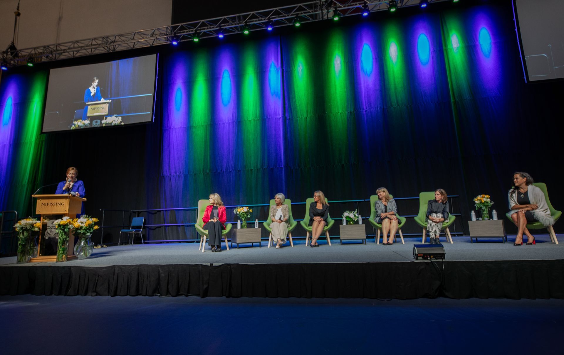 A woman speaking at a podium, panel of women seated on stage with blue, green stage lighting.