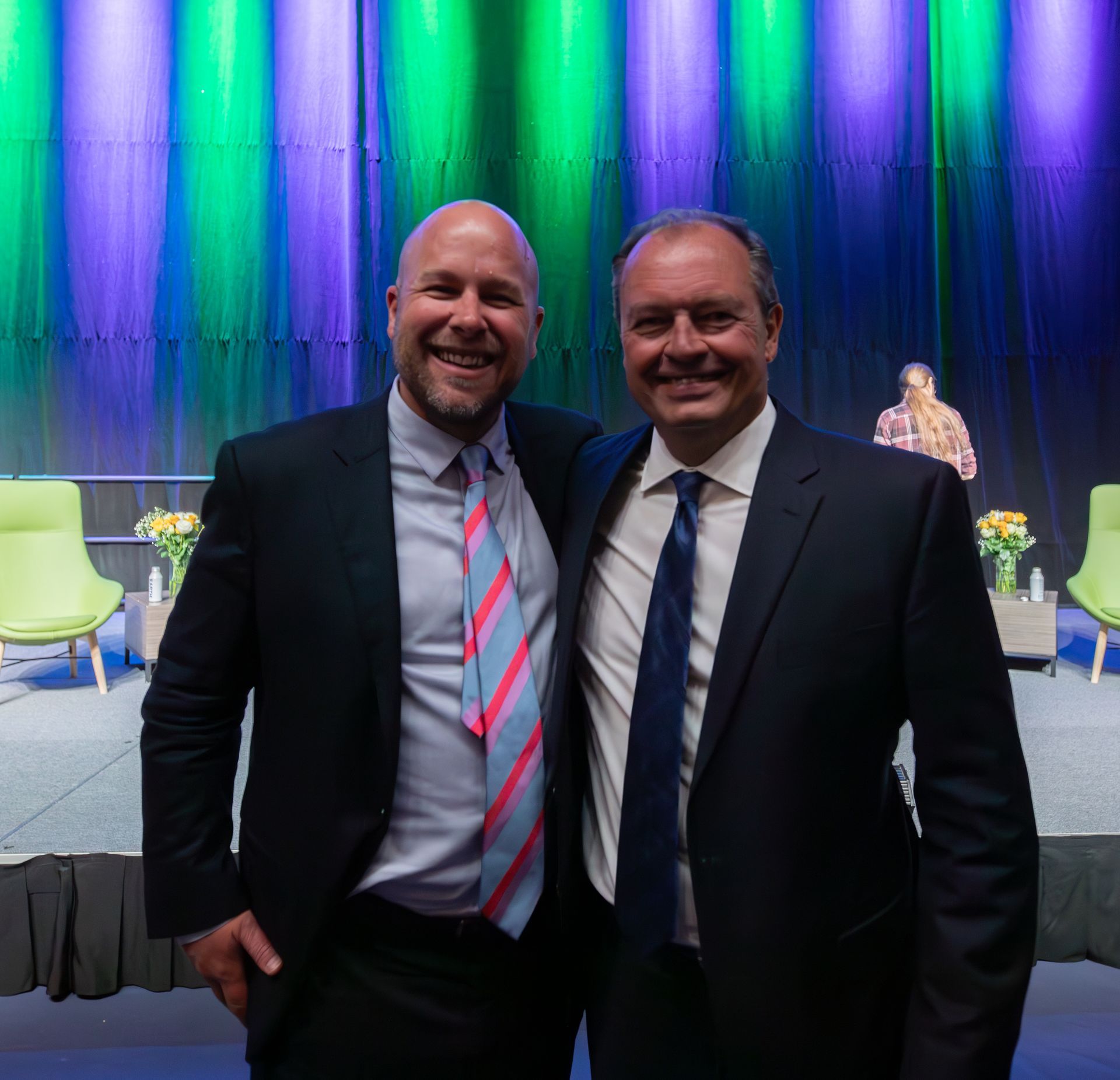 Two men in suits smile, pose on a stage. Green and blue curtains in the background.