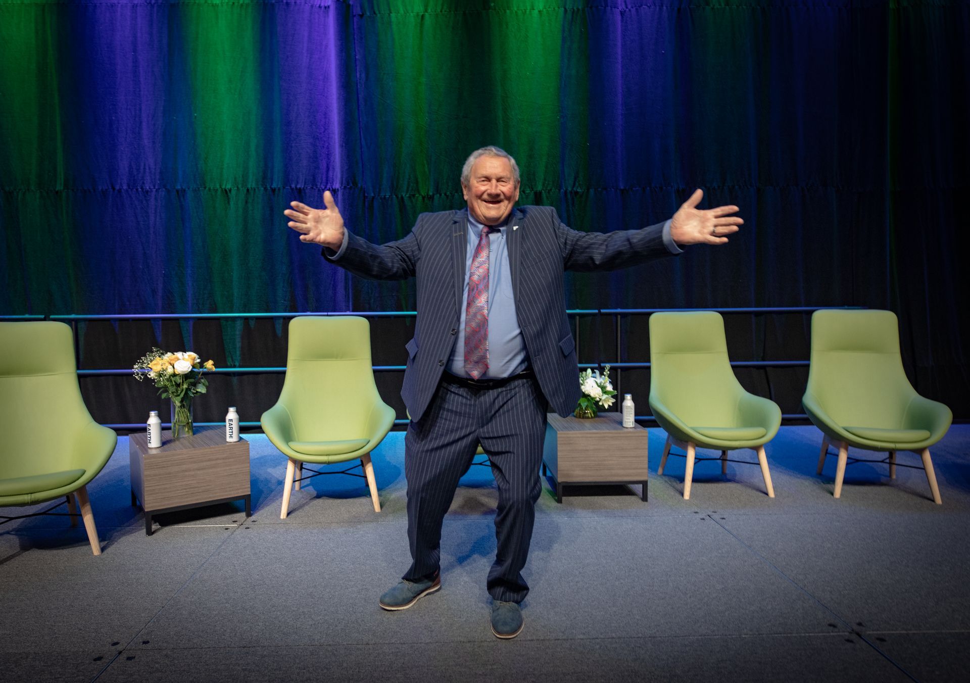 Man in suit with arms outstretched on a stage with green chairs and a colorful background.