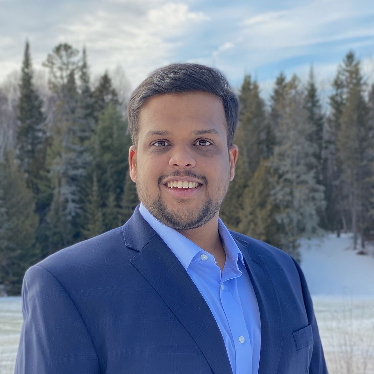 a man in a suit and blue shirt smiles in front of a snowy forest