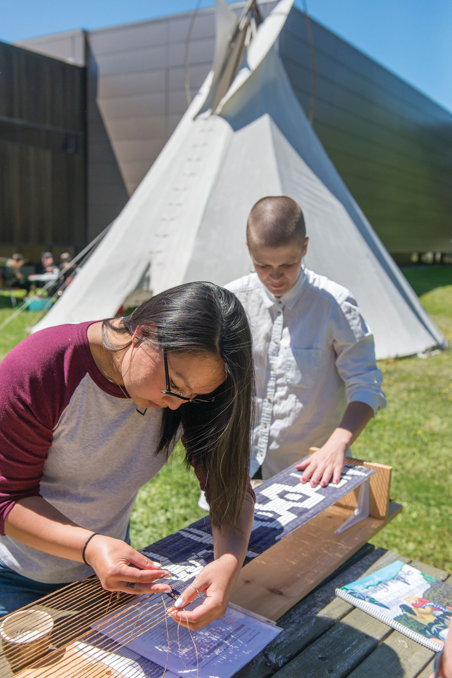 Two people craft outdoors near a tipi. One wears a maroon shirt, beading. The other, in white, assists. Sunny day.