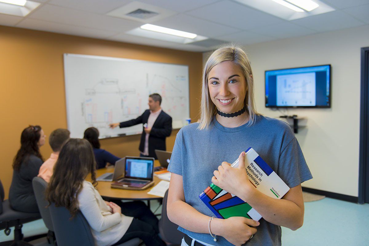 Smiling blonde woman holding a book in a classroom, students and instructor in background.