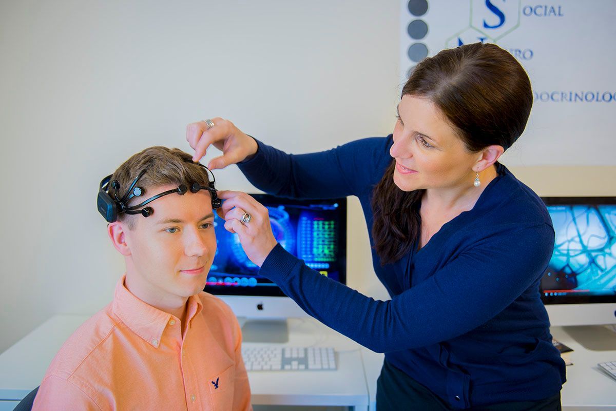 A woman adjusts a brain-scanning device on a man’s head in a research lab with computer screens.