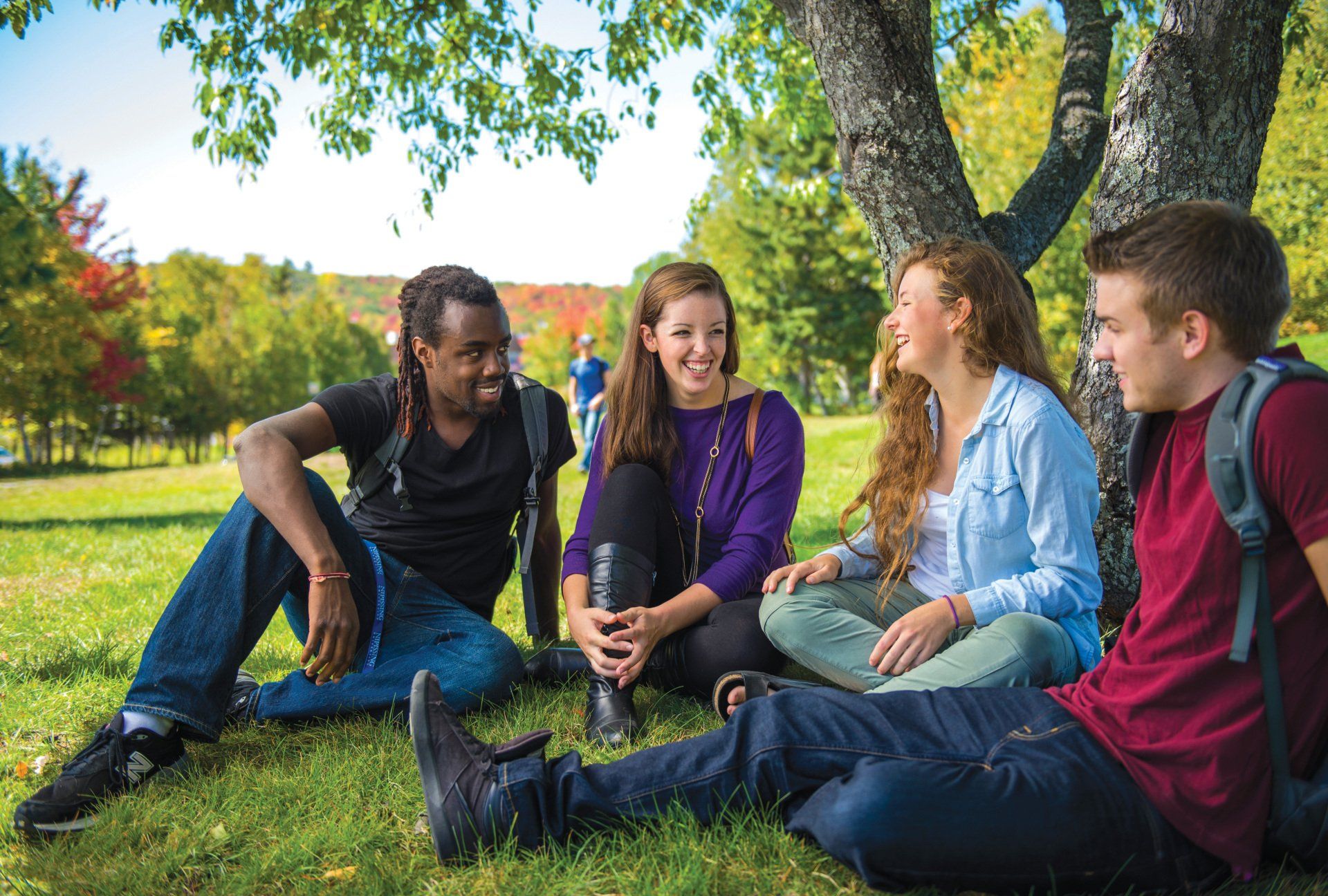 Four college students sit on grass under a tree, smiling and talking. Outdoors in fall, with colorful foliage.