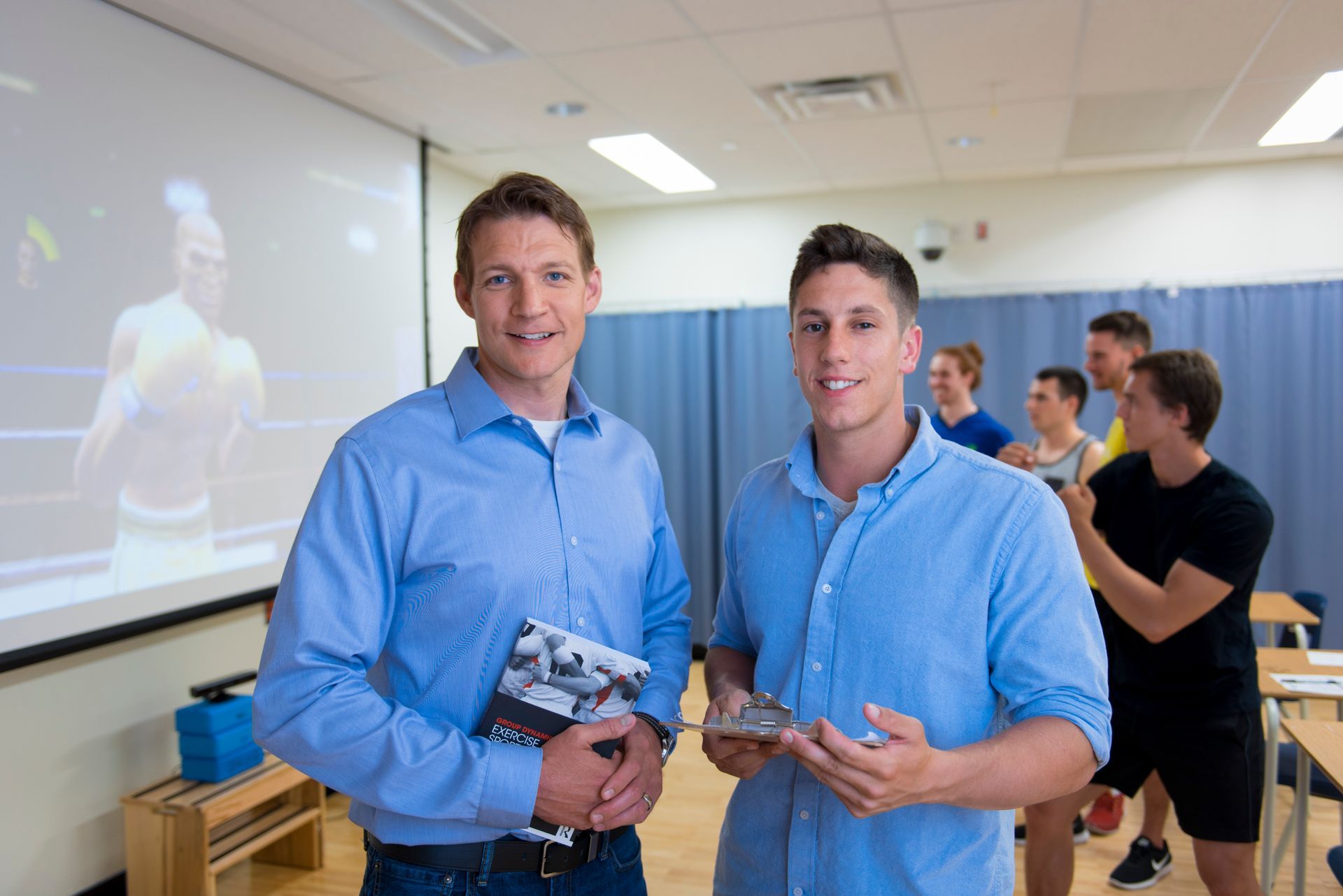 Two men in a classroom, with students in the background.