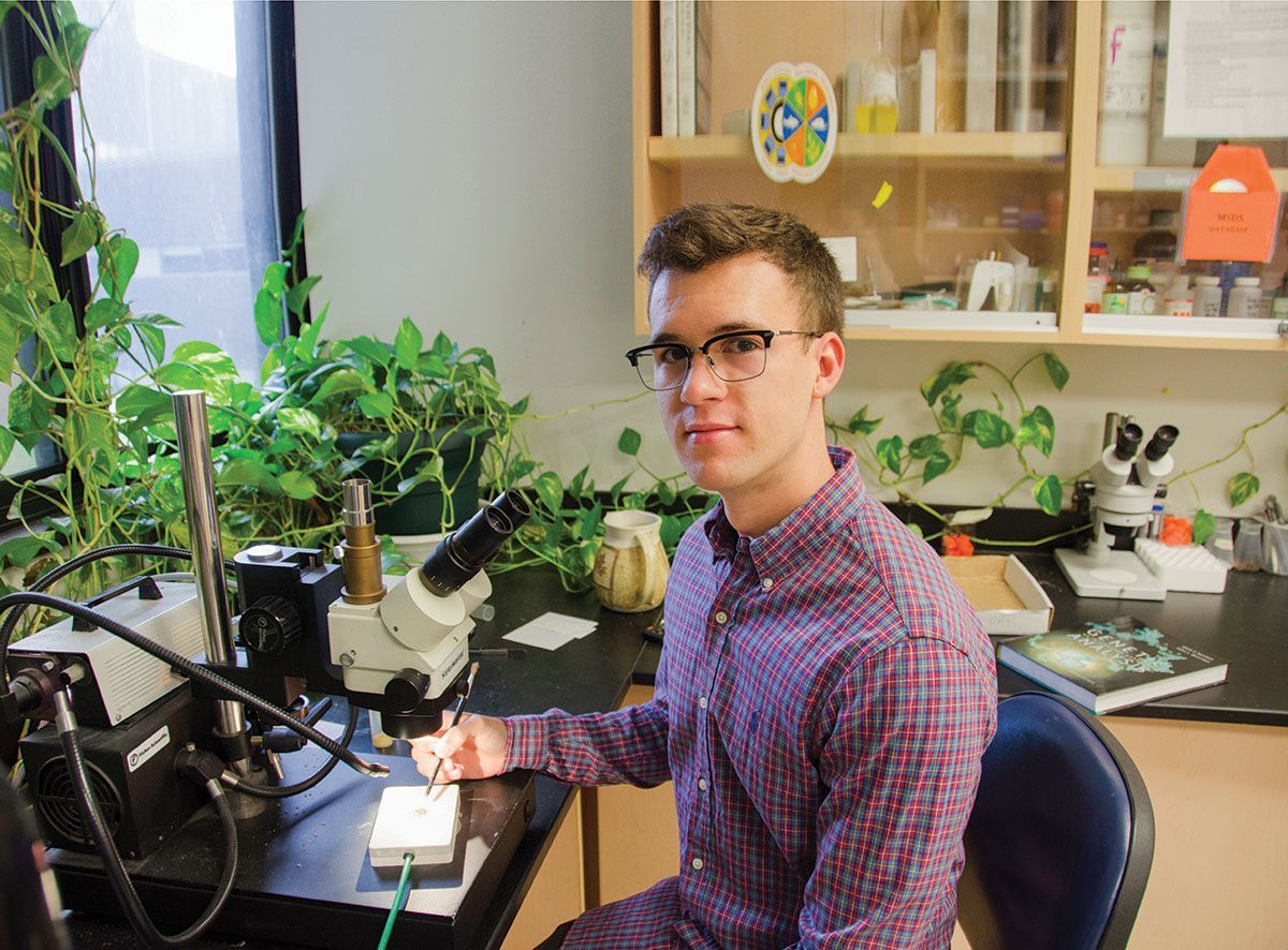 Young man in glasses with a microscope in a lab, surrounded by plants and equipment.