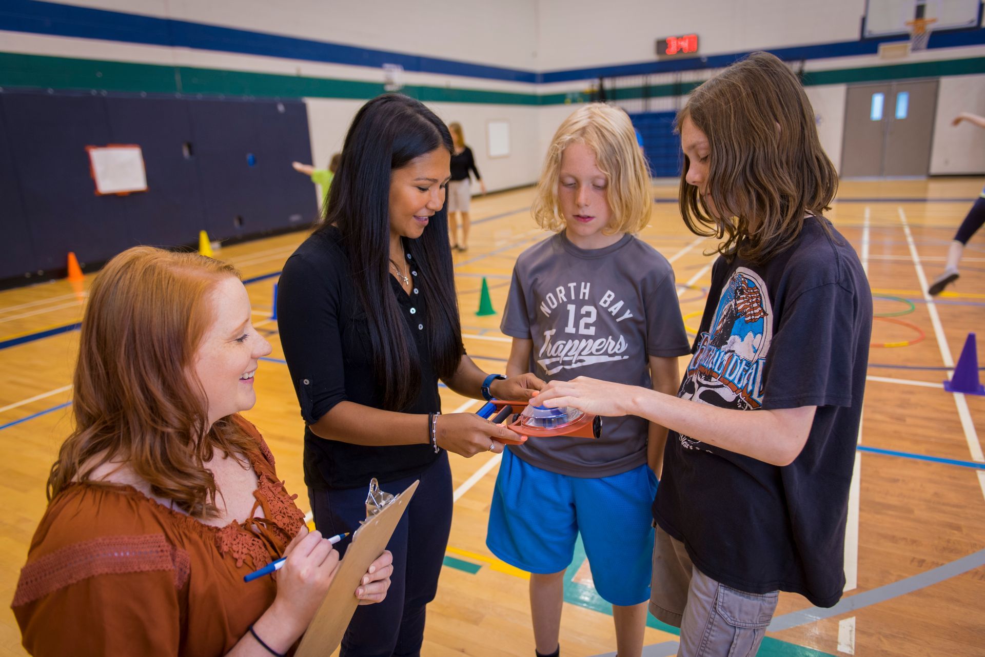 Two women and two children in gym; one woman handing an object to a boy. A woman holds clipboard.