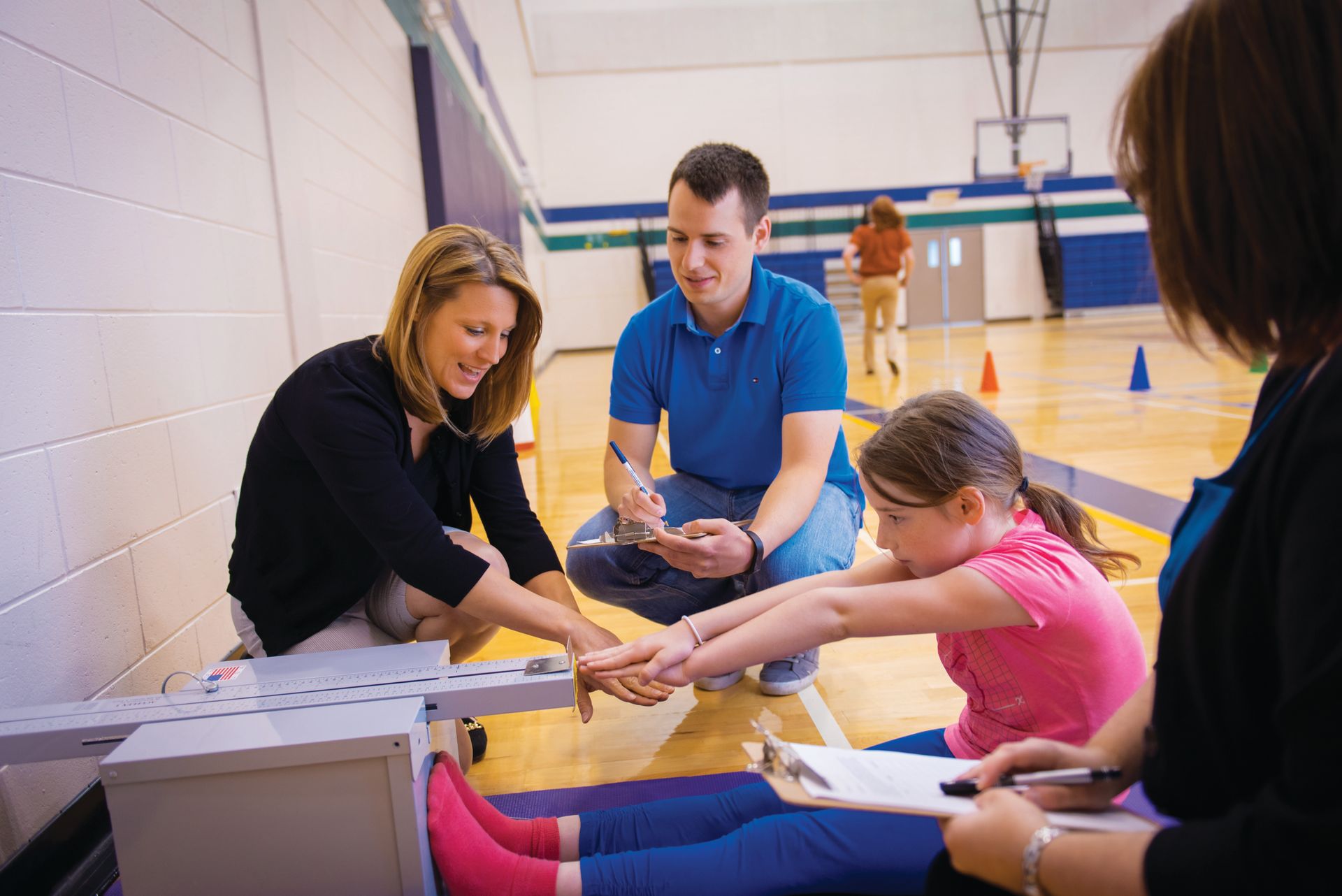 People measure a young girl's flexibility in a gym. A man and woman assist while another woman records the results.