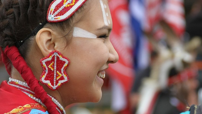 Smiling Indigenous woman in red beaded earrings and headband, wearing face paint, braids with red ties.