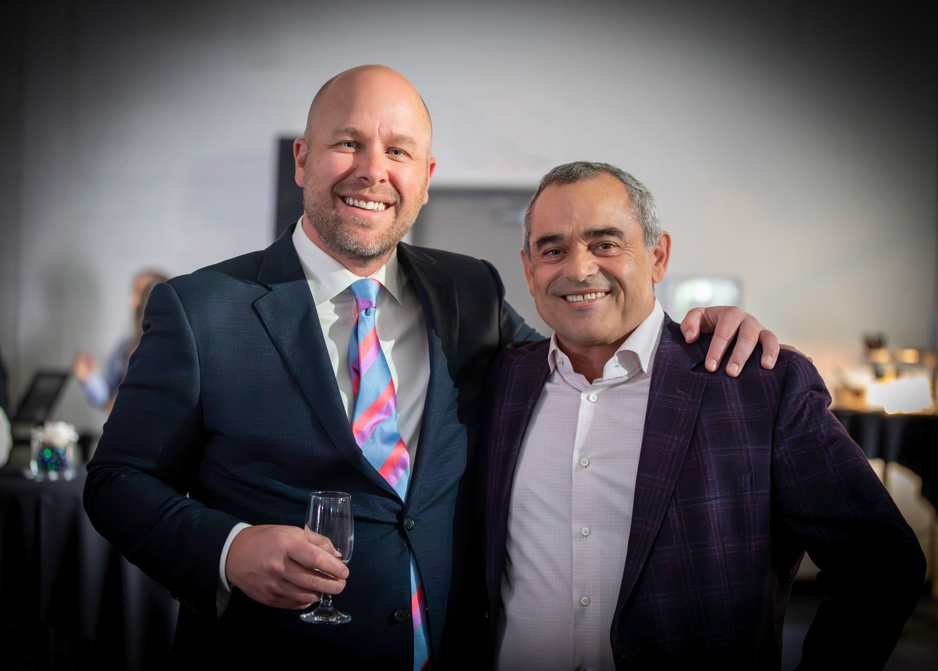 Two men smiling, arms around each other. One in a suit, holding a glass. The other in a purple jacket, indoors.