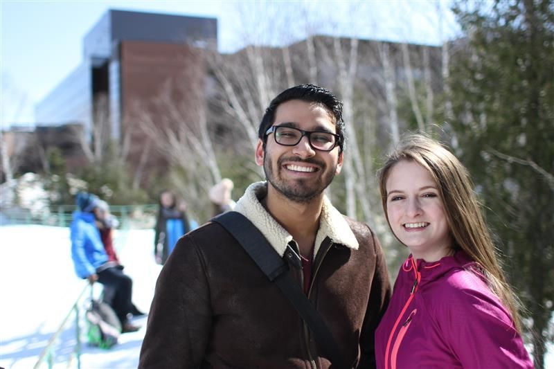Two smiling people outside on a snowy day. Man in glasses and brown jacket, woman in pink jacket. Nipissing University building in the backgrou8nd.